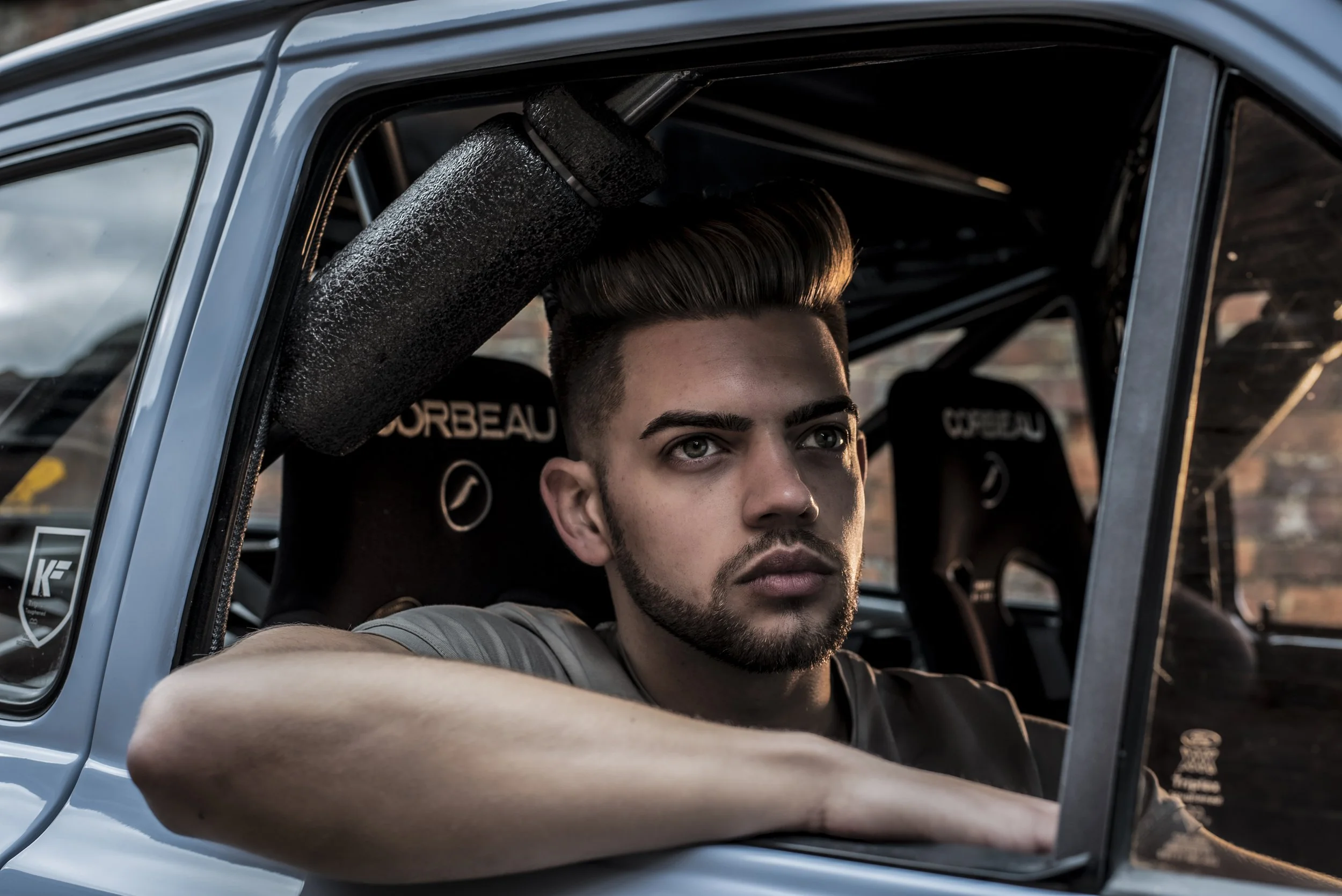 A young man with styled hair and a beard sitting inside a blue car, leaning on the window with his arm, and looking out.