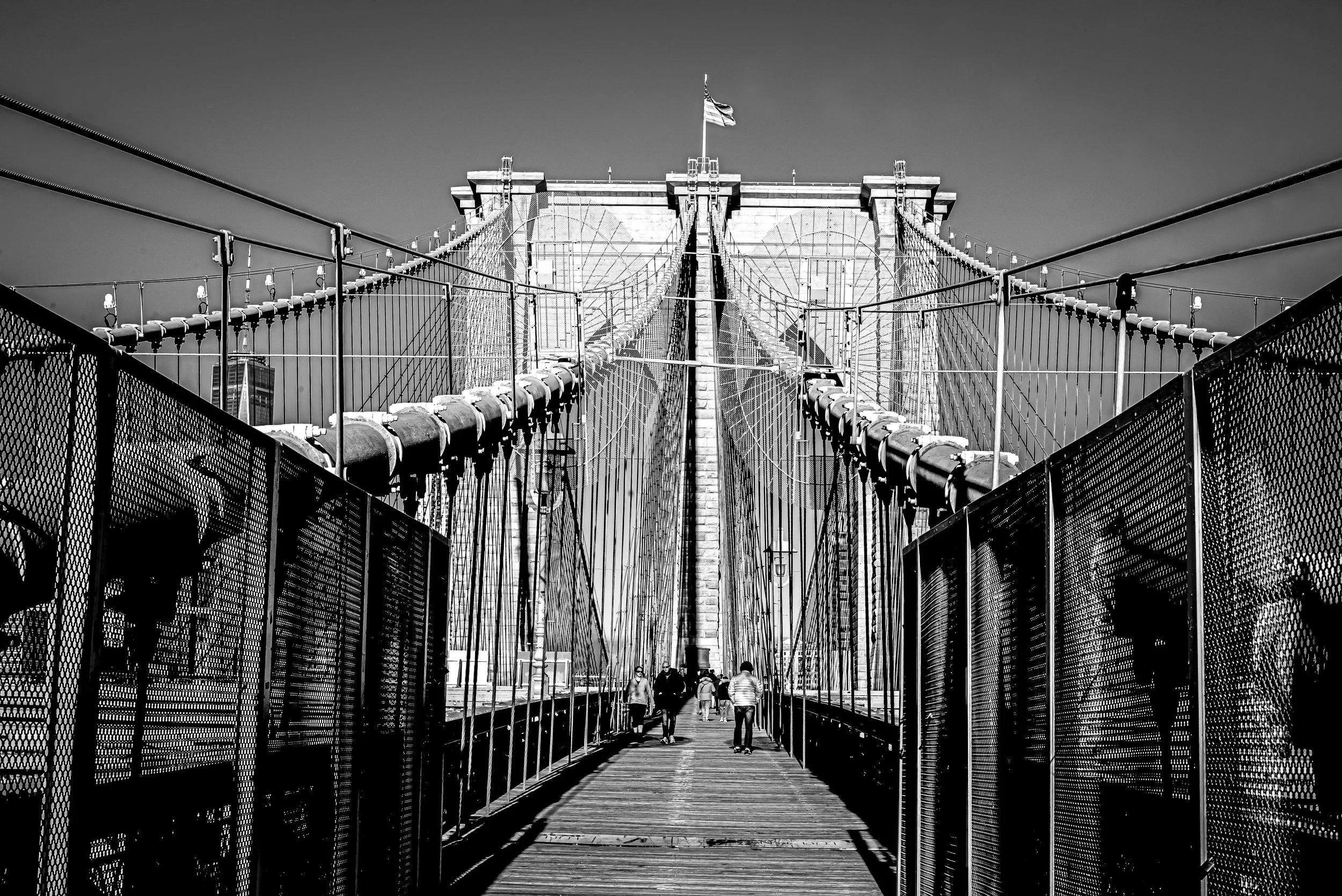 Black and white photo of the Brooklyn Bridge with pedestrians walking on the bridge's walkway.