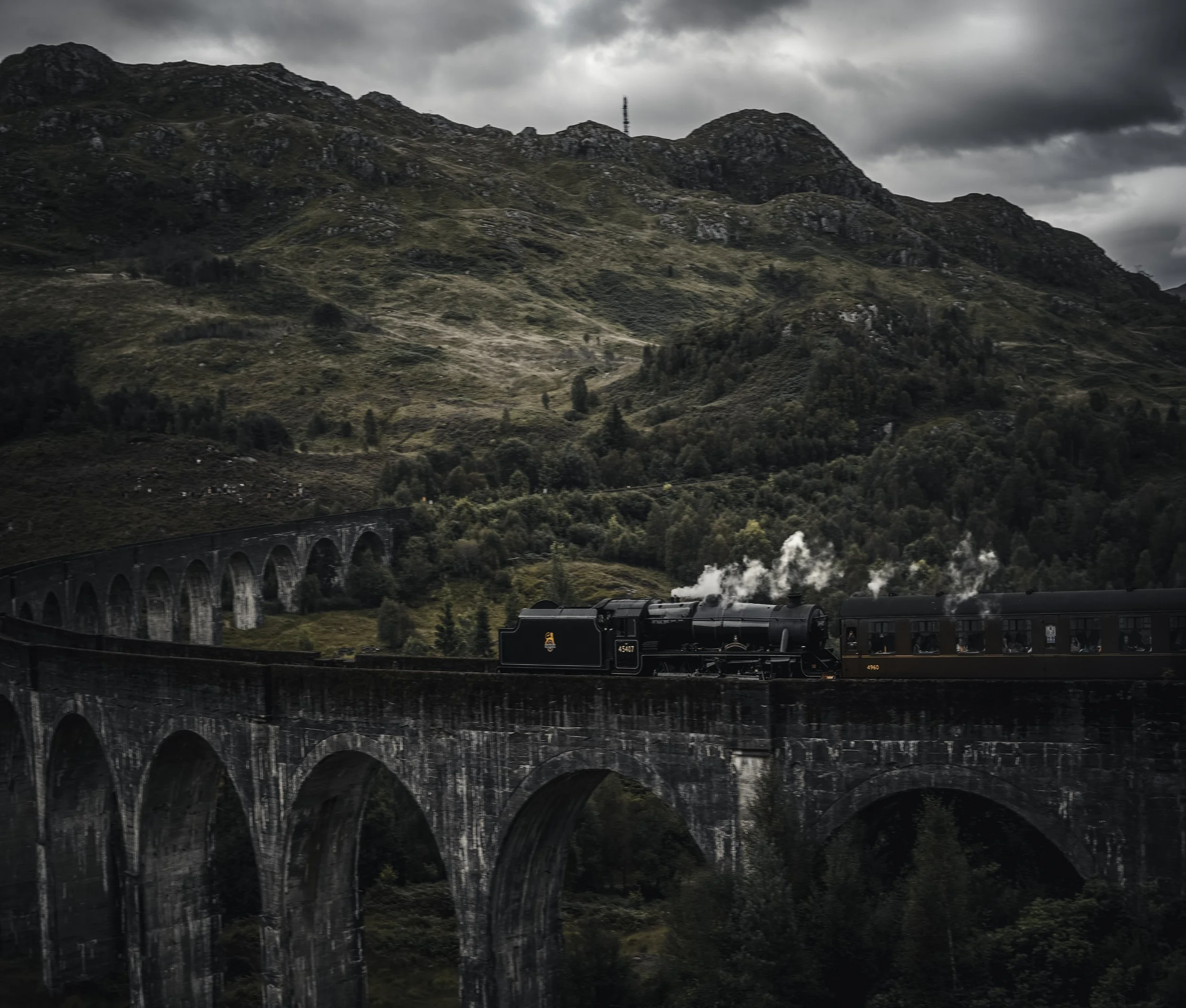 A black steam locomotive train traveling on a curved railway bridge with multiple arches over a lush landscape, set against a mountainous background under an overcast sky.