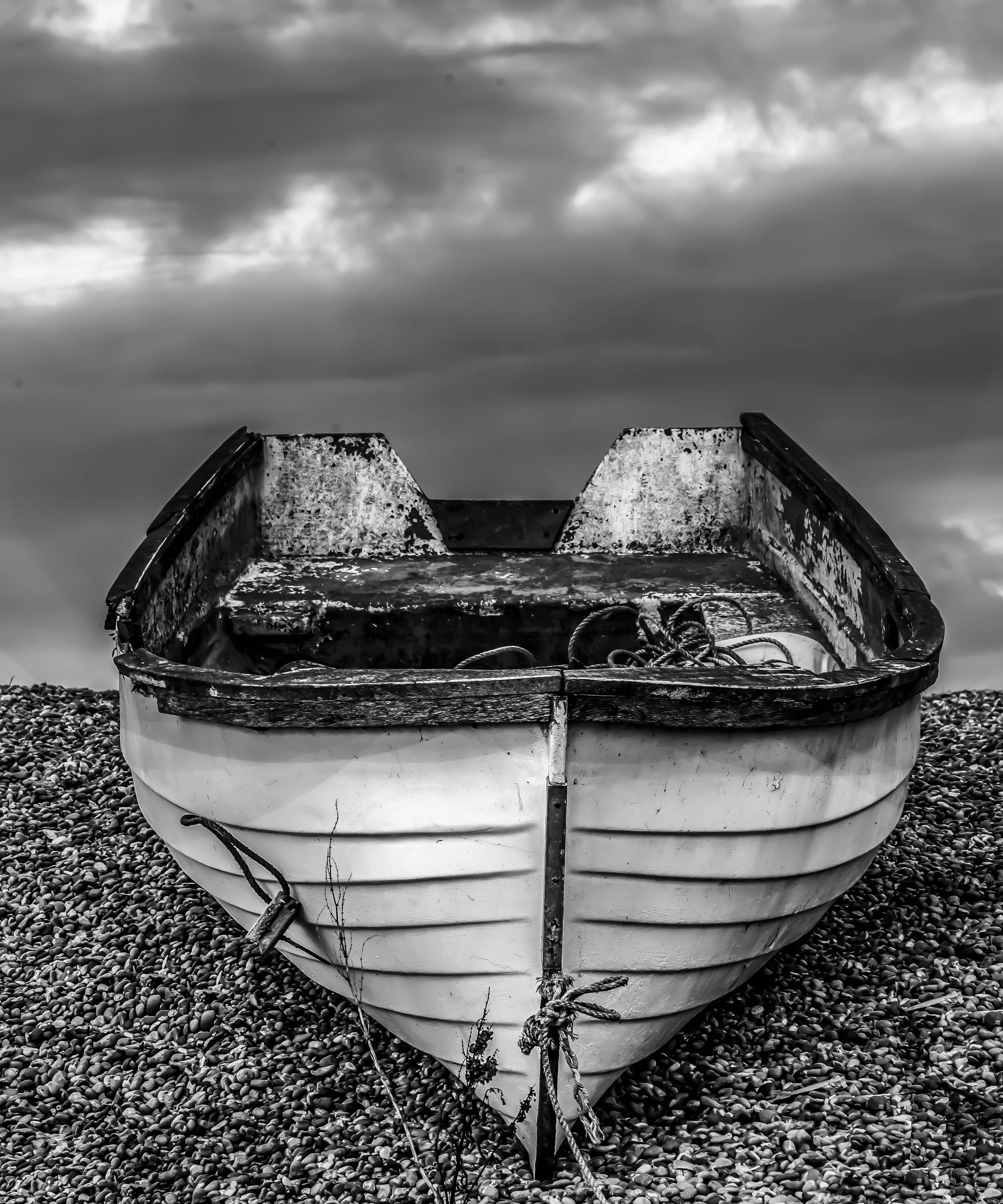 A black and white photo of an old, weathered boat on a pebbled beach, with a cloudy sky in the background.