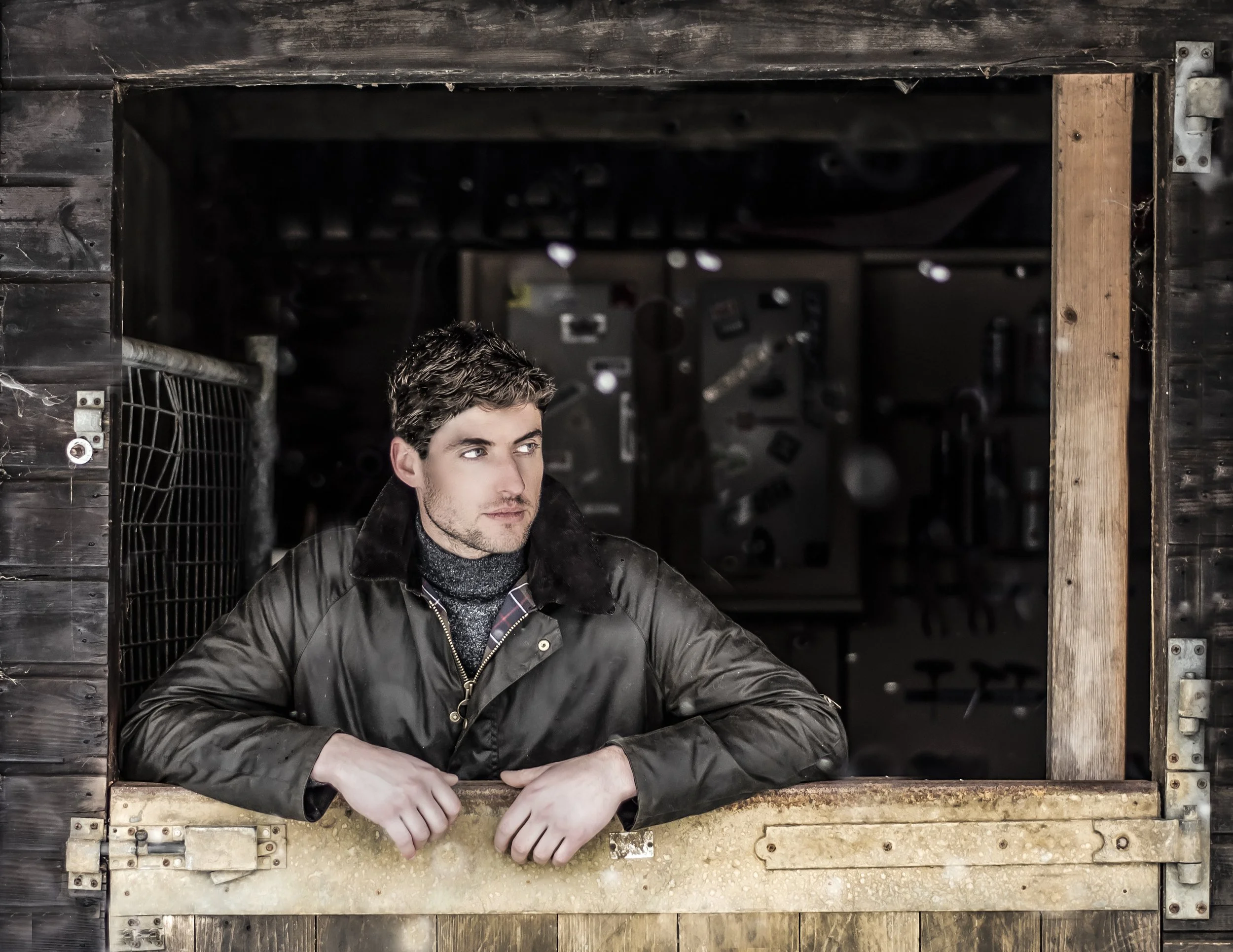 Young man with curly hair and stubble, wearing a black leather jacket, leaning on a wooden railing with his arms crossed, inside a rustic wooden structure with a dark interior and visible metal hardware.
