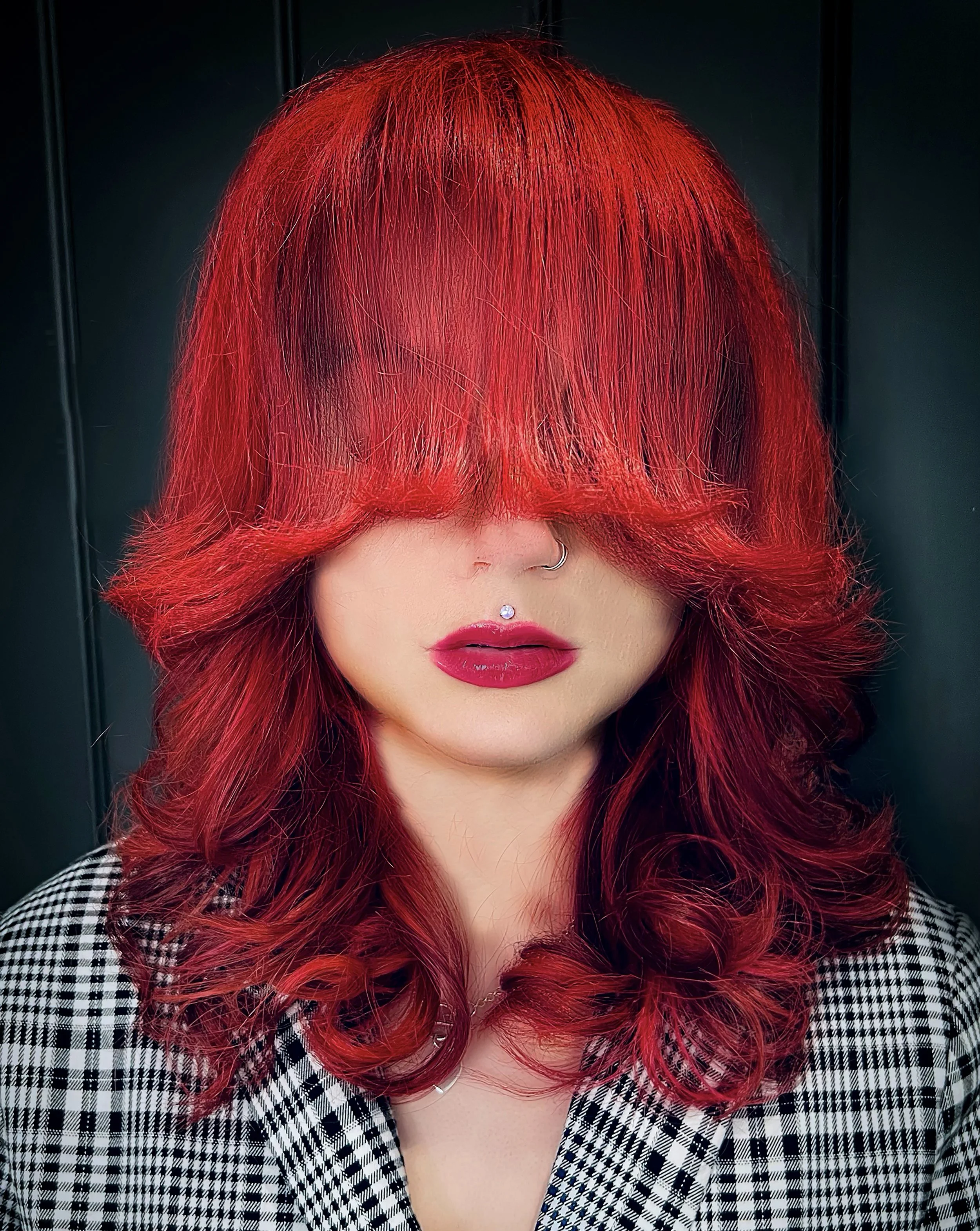 Close-up of a woman with bright red, curly hair covering her eyes, wearing bold red lipstick, a silver nose ring, and a black and white checkered shirt.
