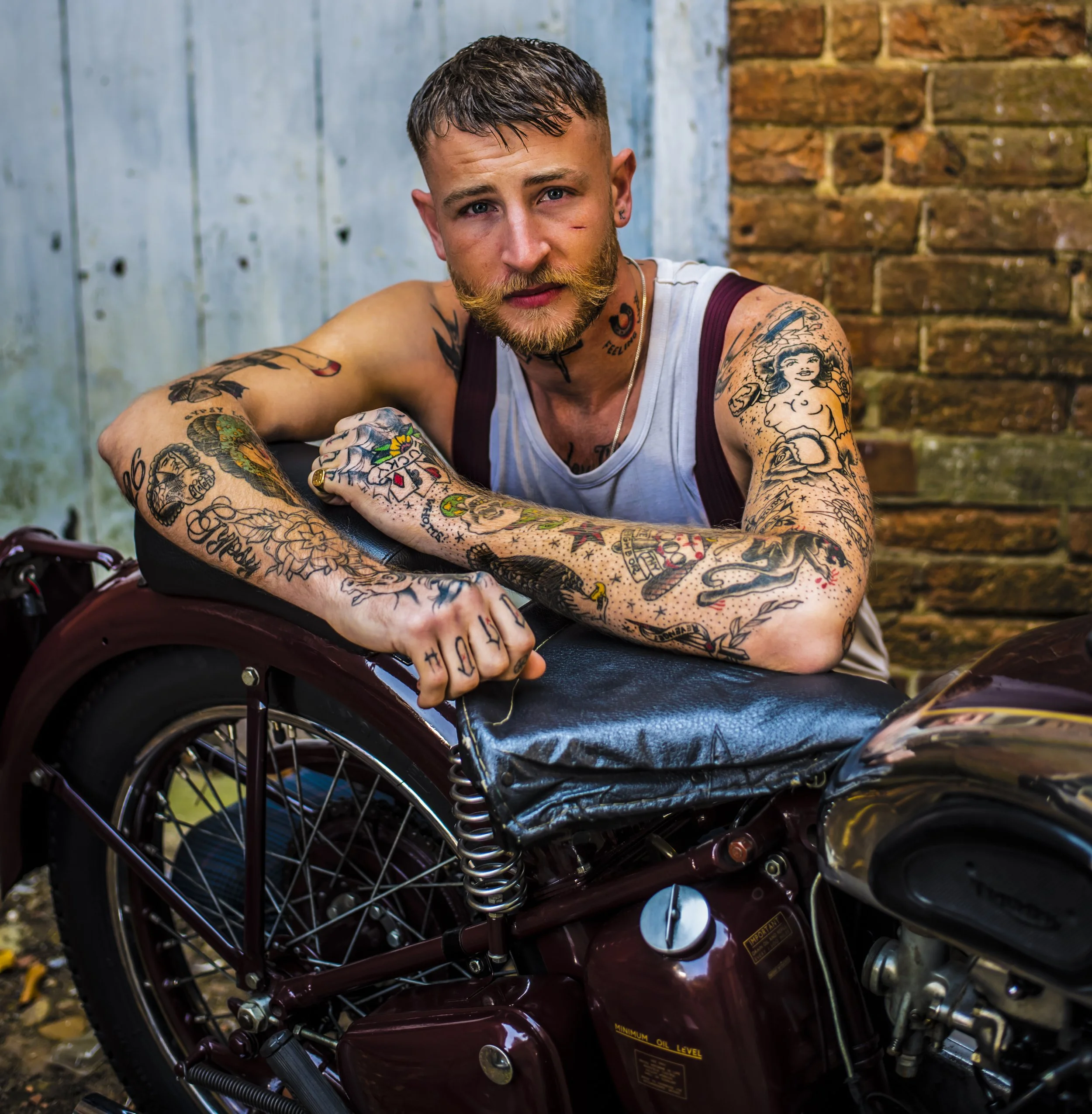 Close-up of a young man with tattoos sitting on a maroon mobility scooter in front of a brick wall and wooden panel background, looking at the camera with blue eyes and a slight smile.