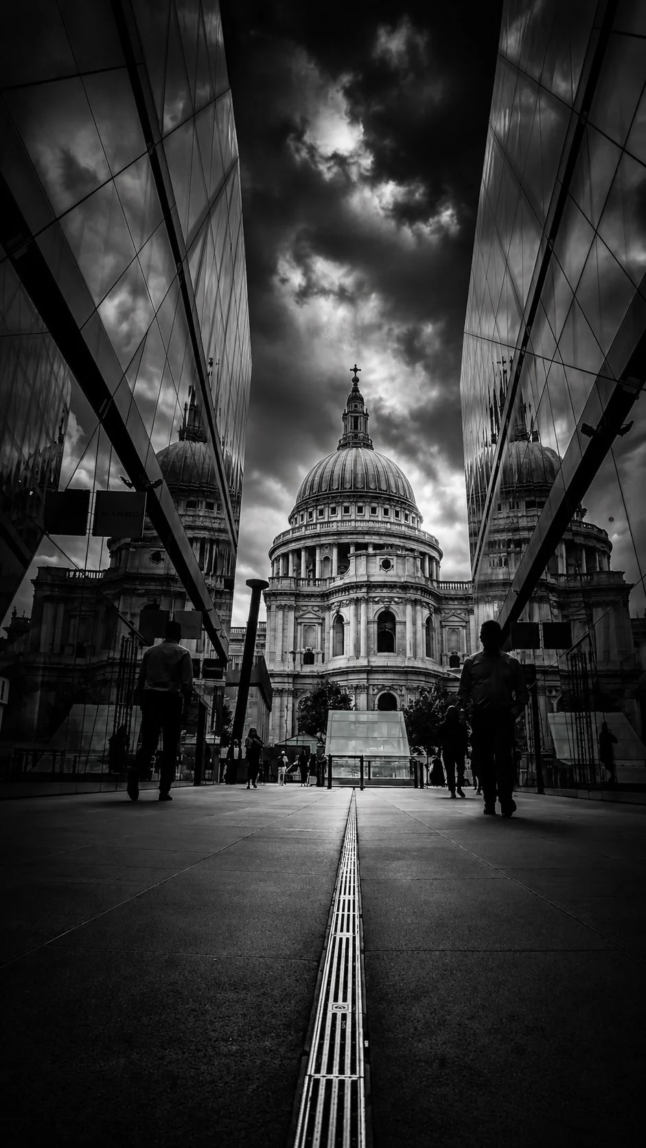 Black and white photo of St. Paul's Cathedral in London, captured between modern glass buildings with a cloudy sky overhead. Several people walk on the pavement.