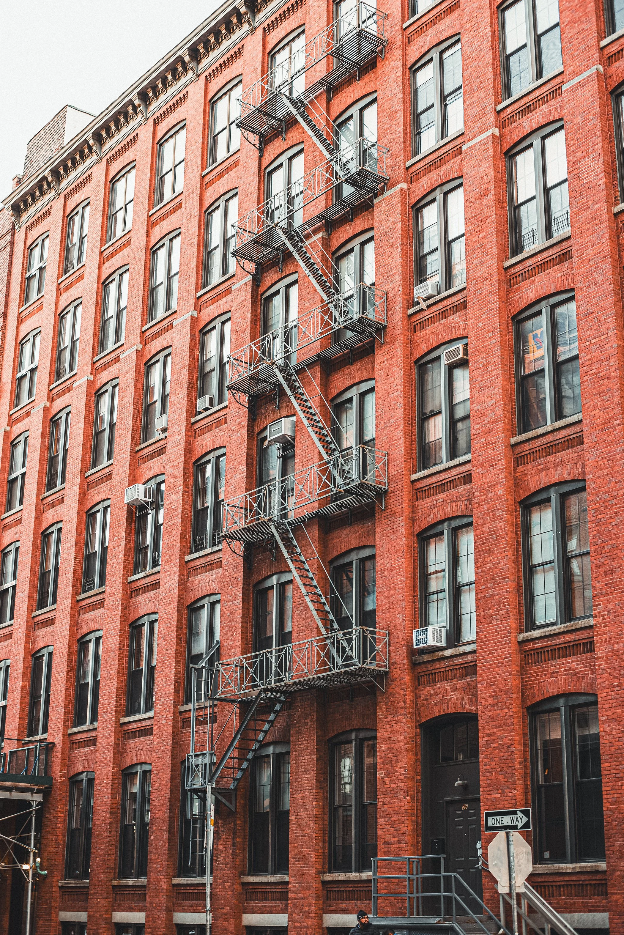 A red brick multi-story building with large arched windows and fire escapes on the front. Air conditioning units are attached to some windows, and there are street signs and a person at the bottom.