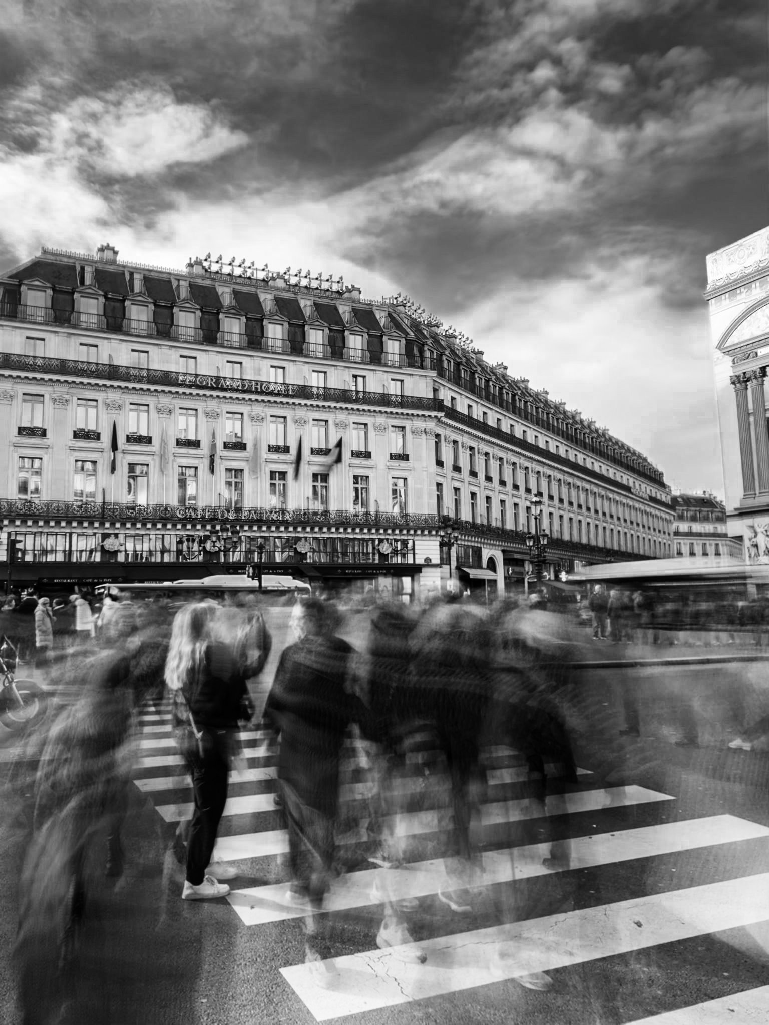 Black and white photo of a city street with a crosswalk, blurred pedestrians, historic building with ornate architecture, flag, and part of an arch monument under a cloudy sky.