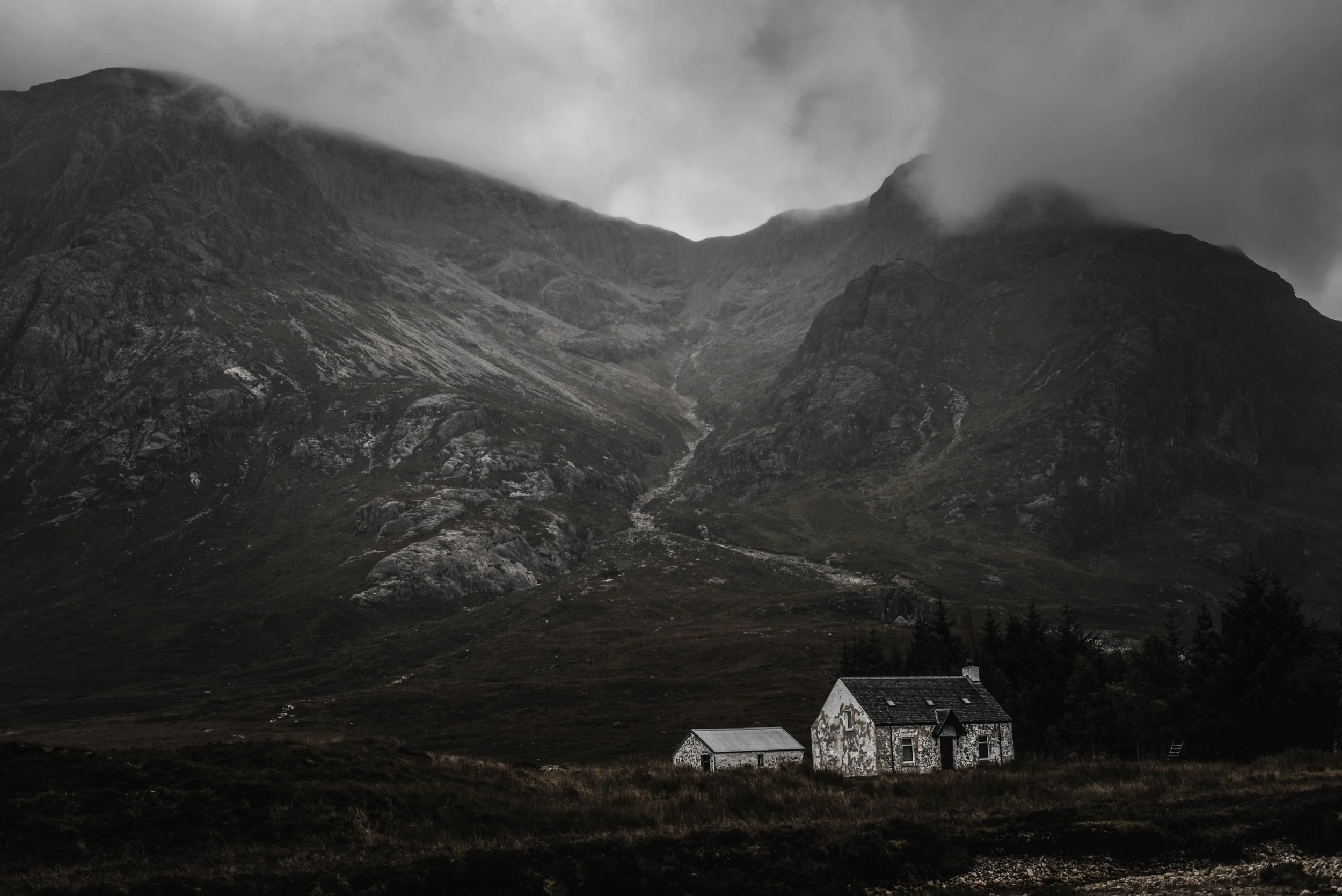 A landscape of dark mountains with cloudy skies, a small white house with a dark roof in the lower right corner, and a few other small buildings nearby.