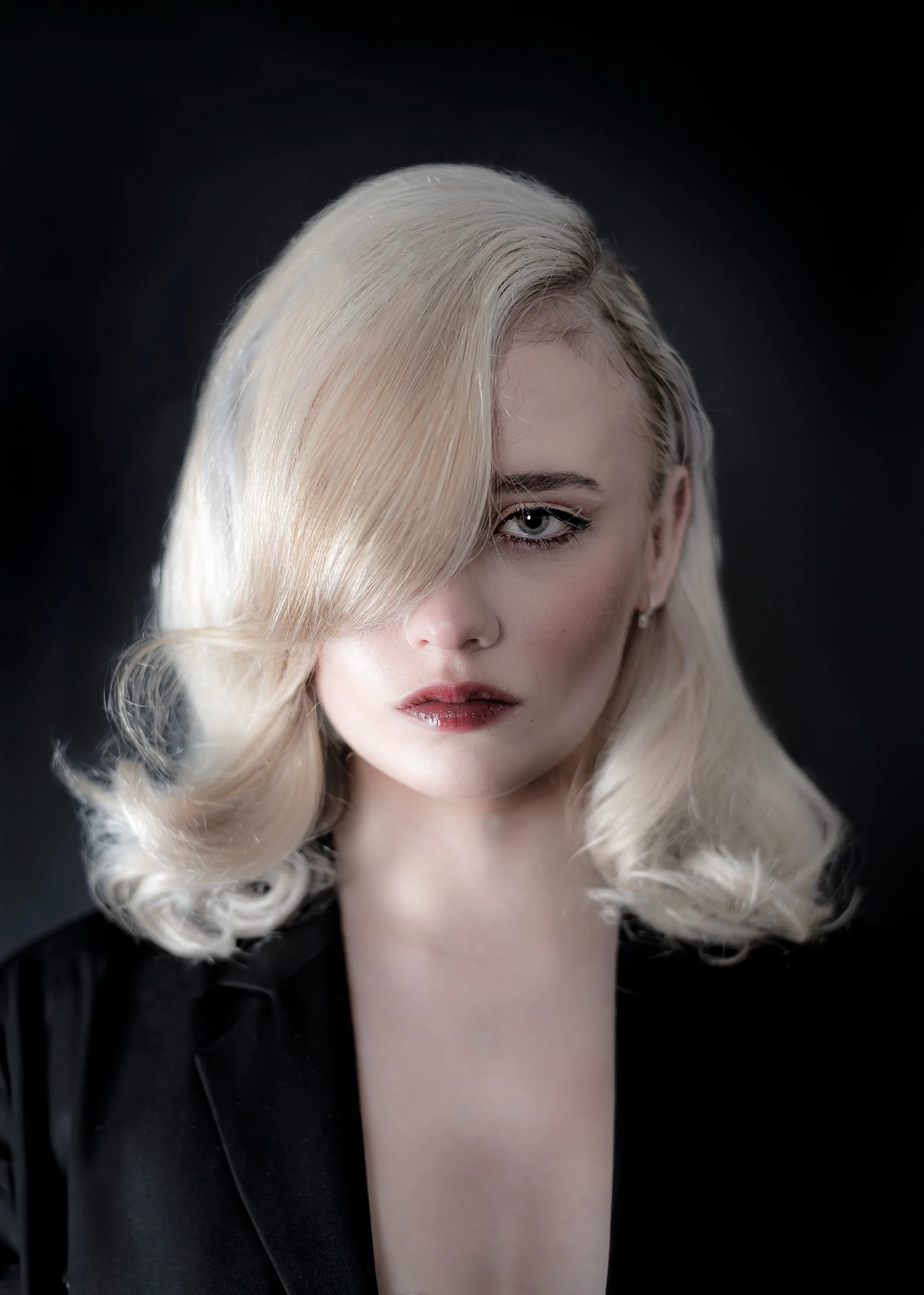 A woman with platinum blonde hair styled in vintage glam waves, with one side covering part of her face, against a dark background.