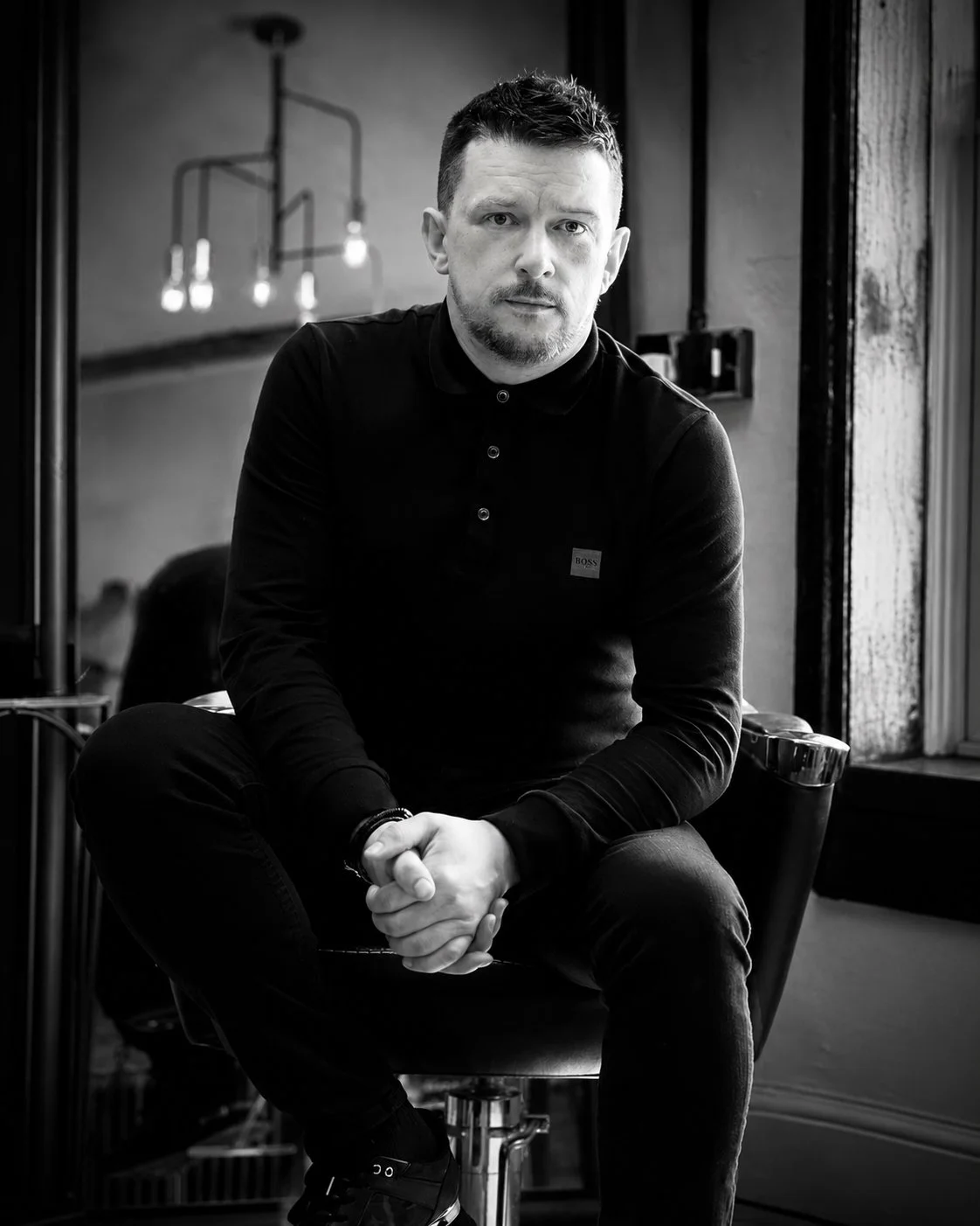 A black-and-white photo of a man sitting on a chair in a room with a large window. He has short dark hair, a beard, and is wearing a dark polo shirt and dark pants. He is looking directly at the camera with a serious expression, sitting with his hands clasped. In the background, there are some pendant lights hanging from the ceiling and a mirror reflecting part of the room.