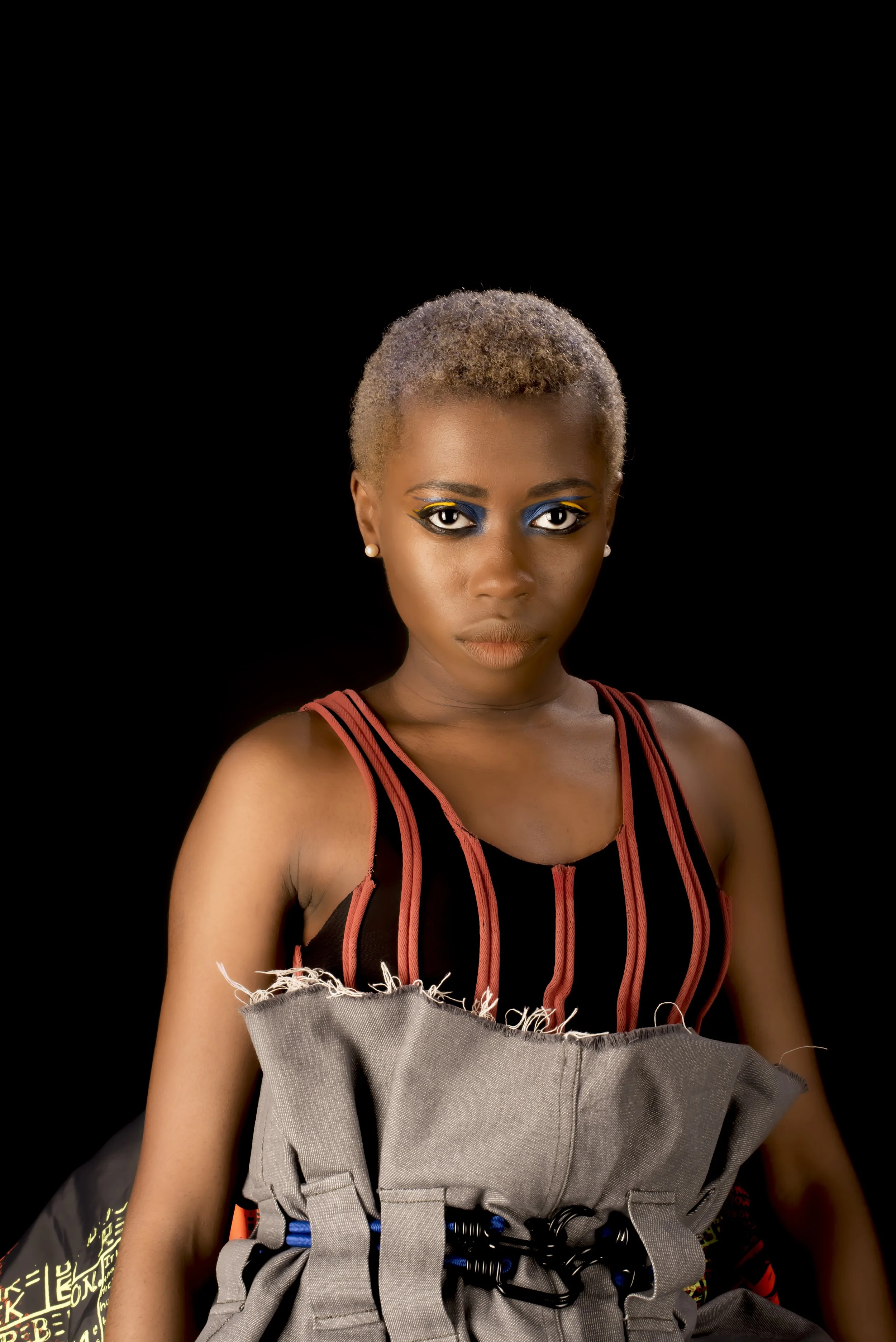 Portrait of a woman with short blond hair, bold blue and yellow eye makeup, wearing a black top with orange and pink vertical stripes and a gray utility vest, against a black background.