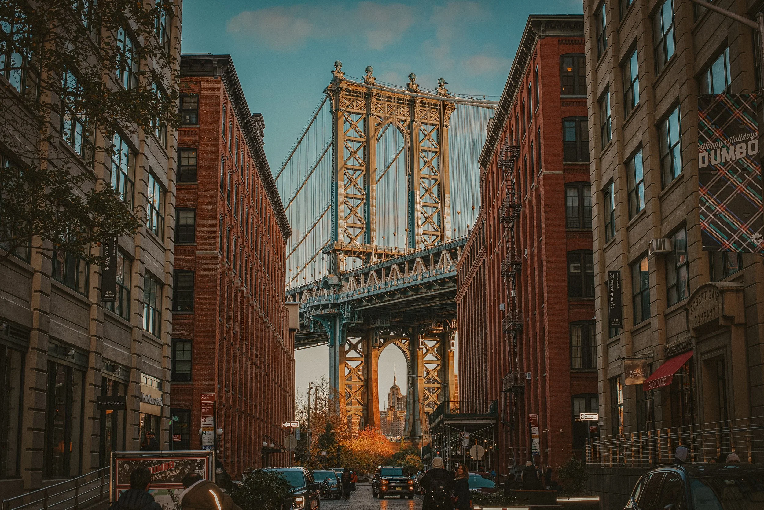 View of Manhattan Bridge seen through a city street lined with brick buildings on both sides, with cars and pedestrians in the foreground, and a clear blue sky in the background.