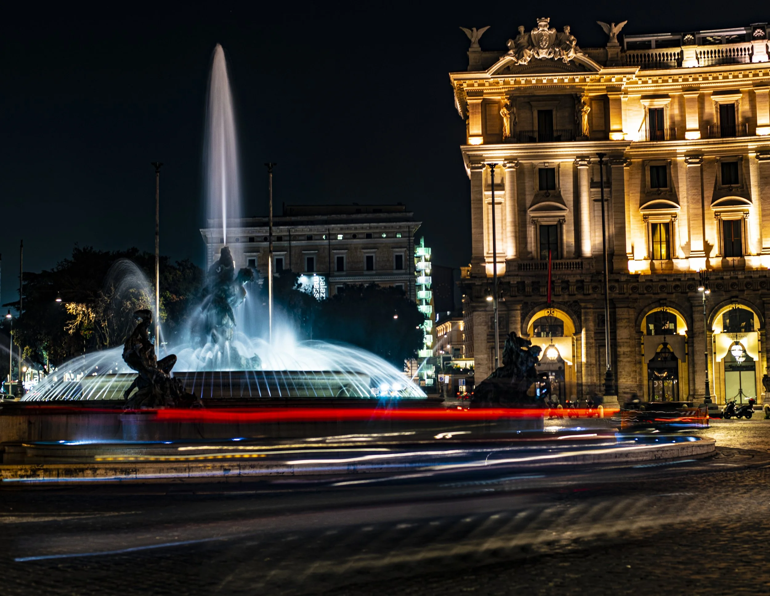 Nighttime scene featuring a lit historic building with statues on the roof, a fountain with sculptures in the foreground, and a water display with streaks from moving vehicles.