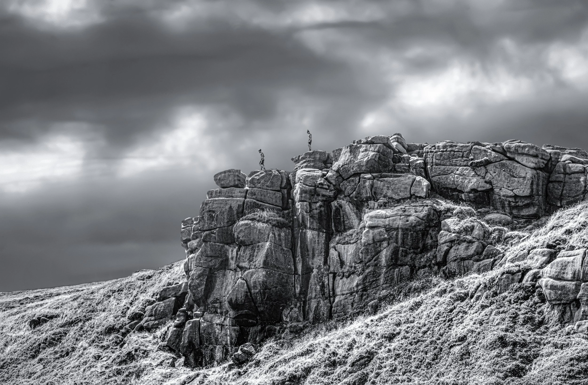 Black and white photograph of two individuals mountain climbing on a steep rocky cliff under dark, cloudy skies.