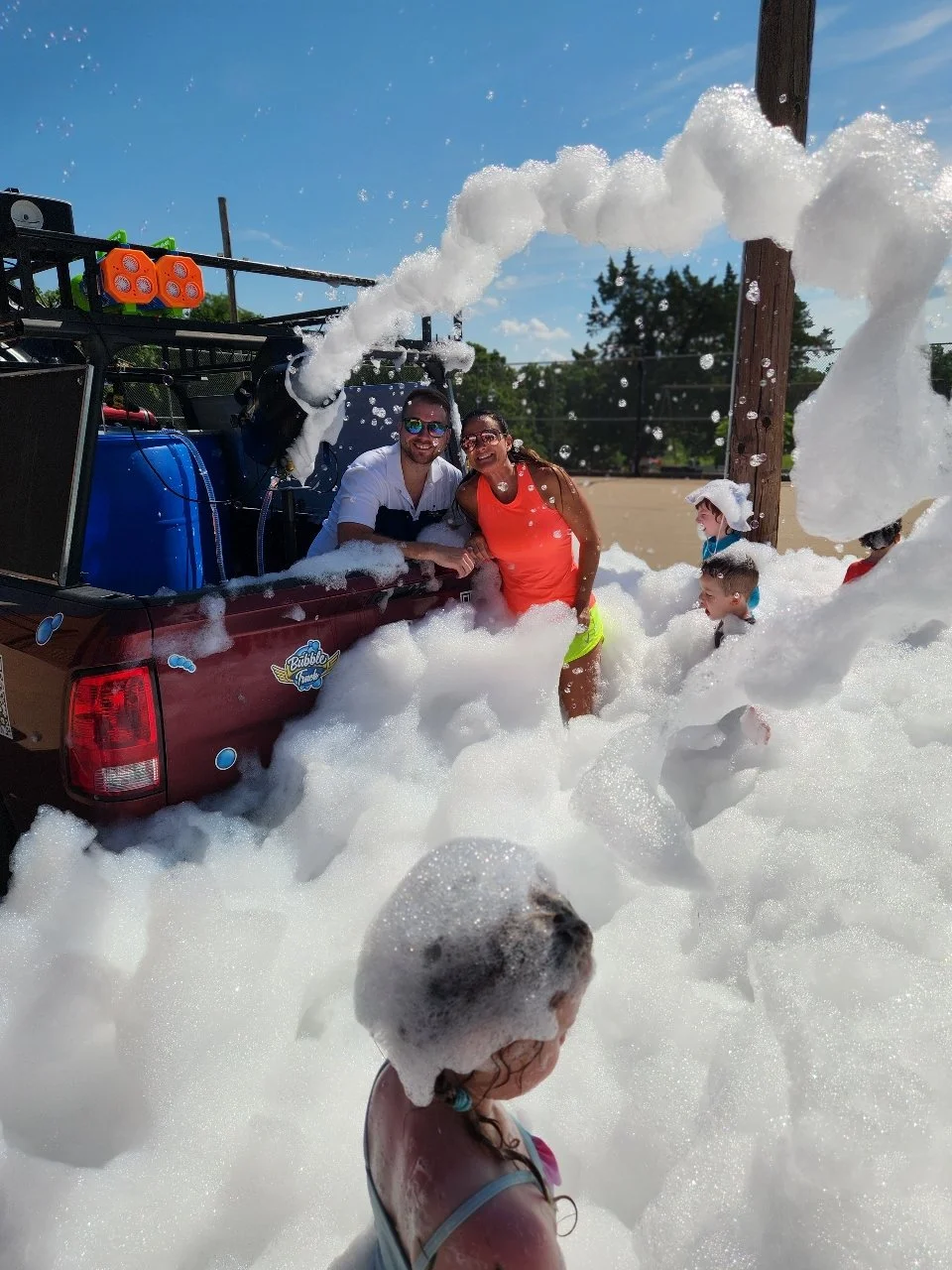 People having fun in a foam party outdoors on a sunny day, with a truck releasing foam, children and adults enjoying the foam, some wearing swimsuits and hats.