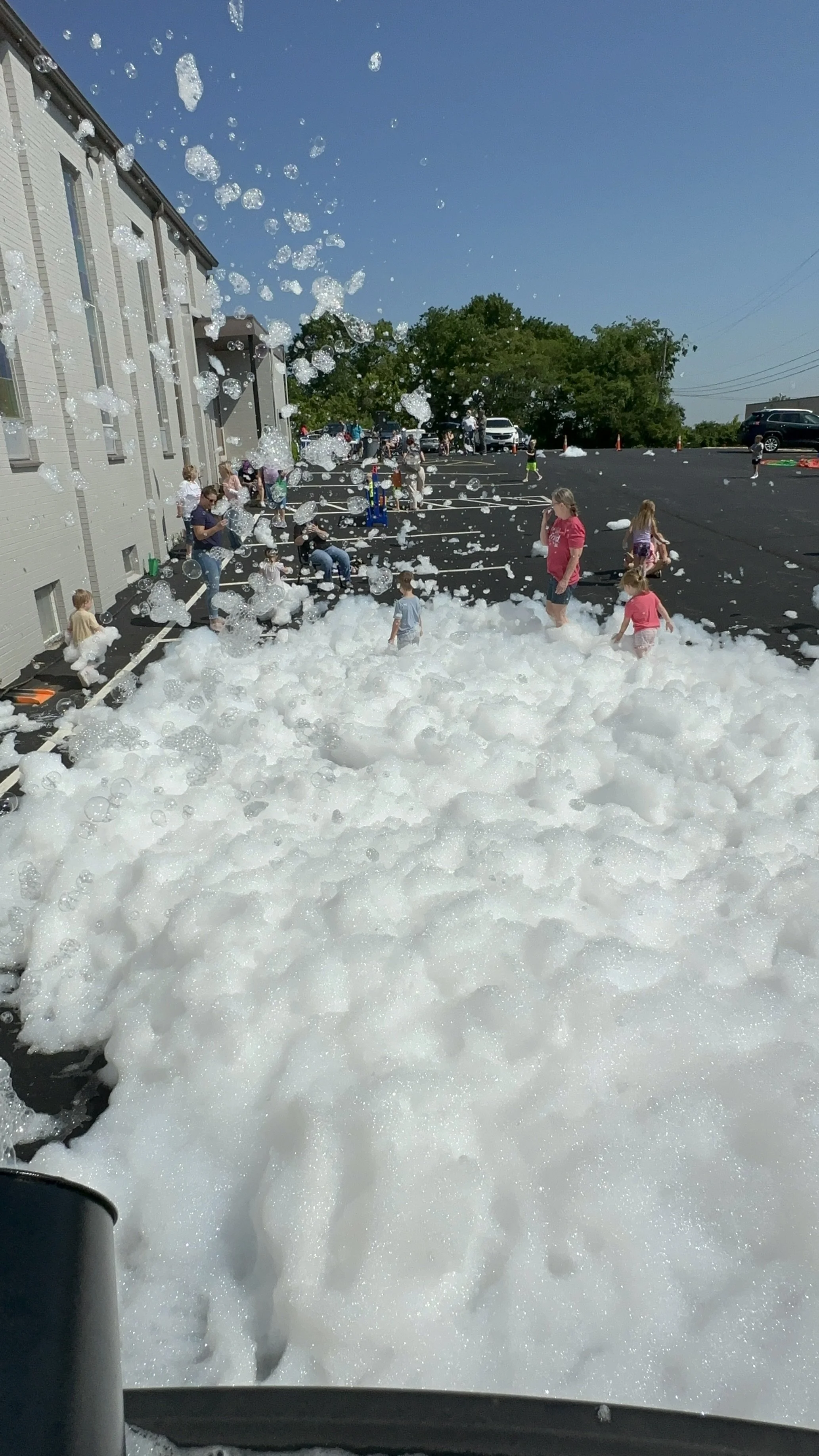 Children and adults playing in a large foam or soap bubble foam outside a building on a sunny day.
