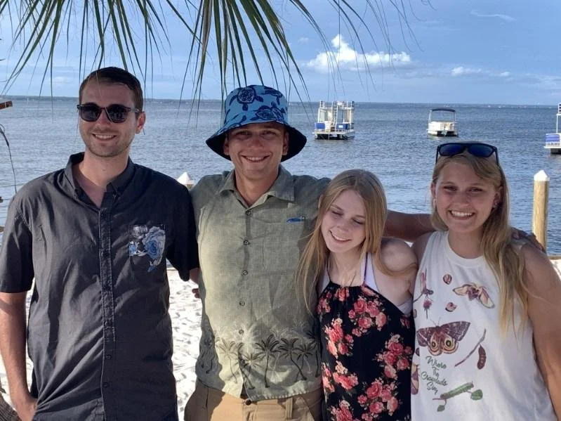 Four people posing on a beach with boats on the water in the background, under a palm tree.