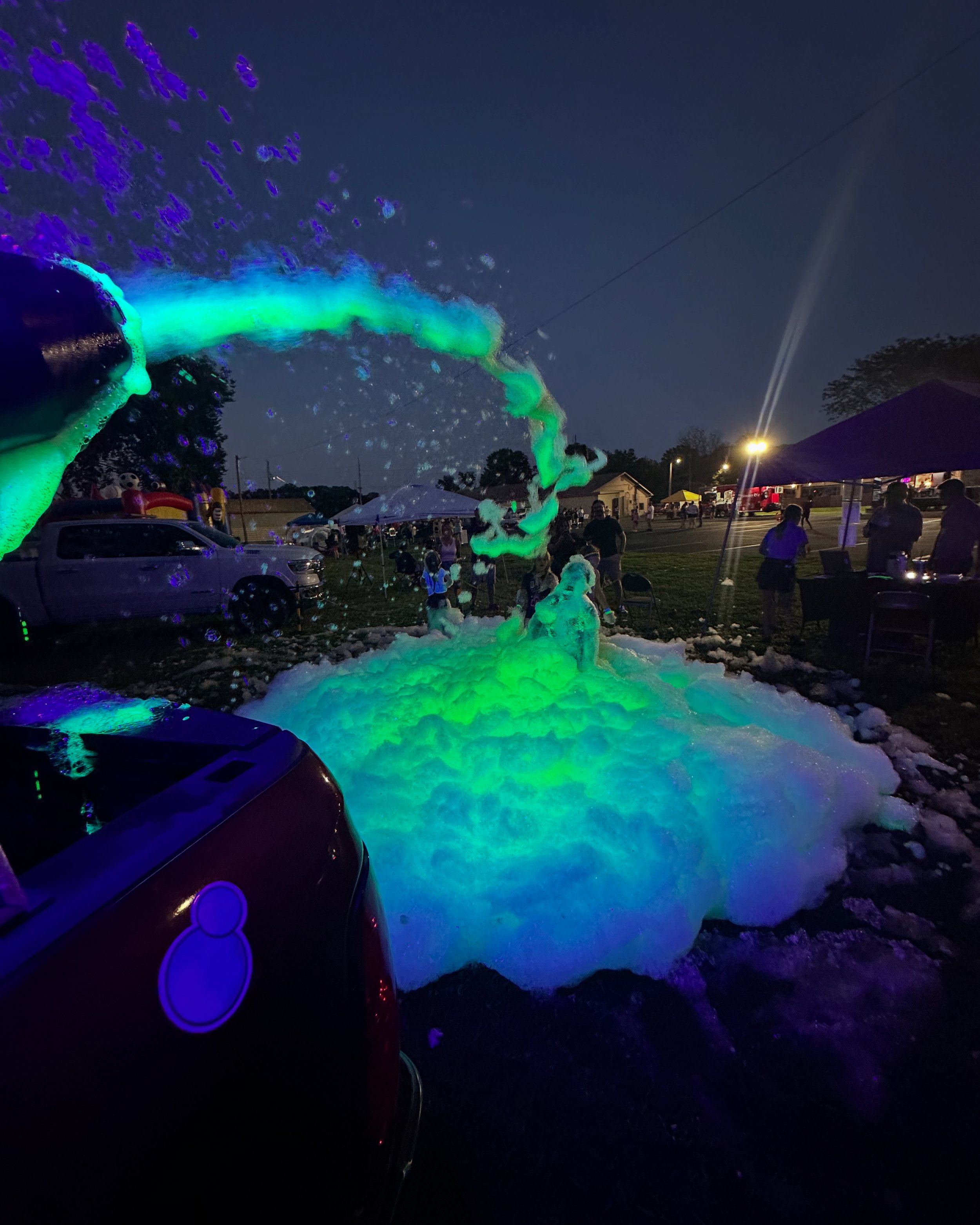Night scene with a person creating glowing foam with a foam machine, colorful foam emitting blue and green glow. People gathered around, tents, parking lot, and trees in the background.
