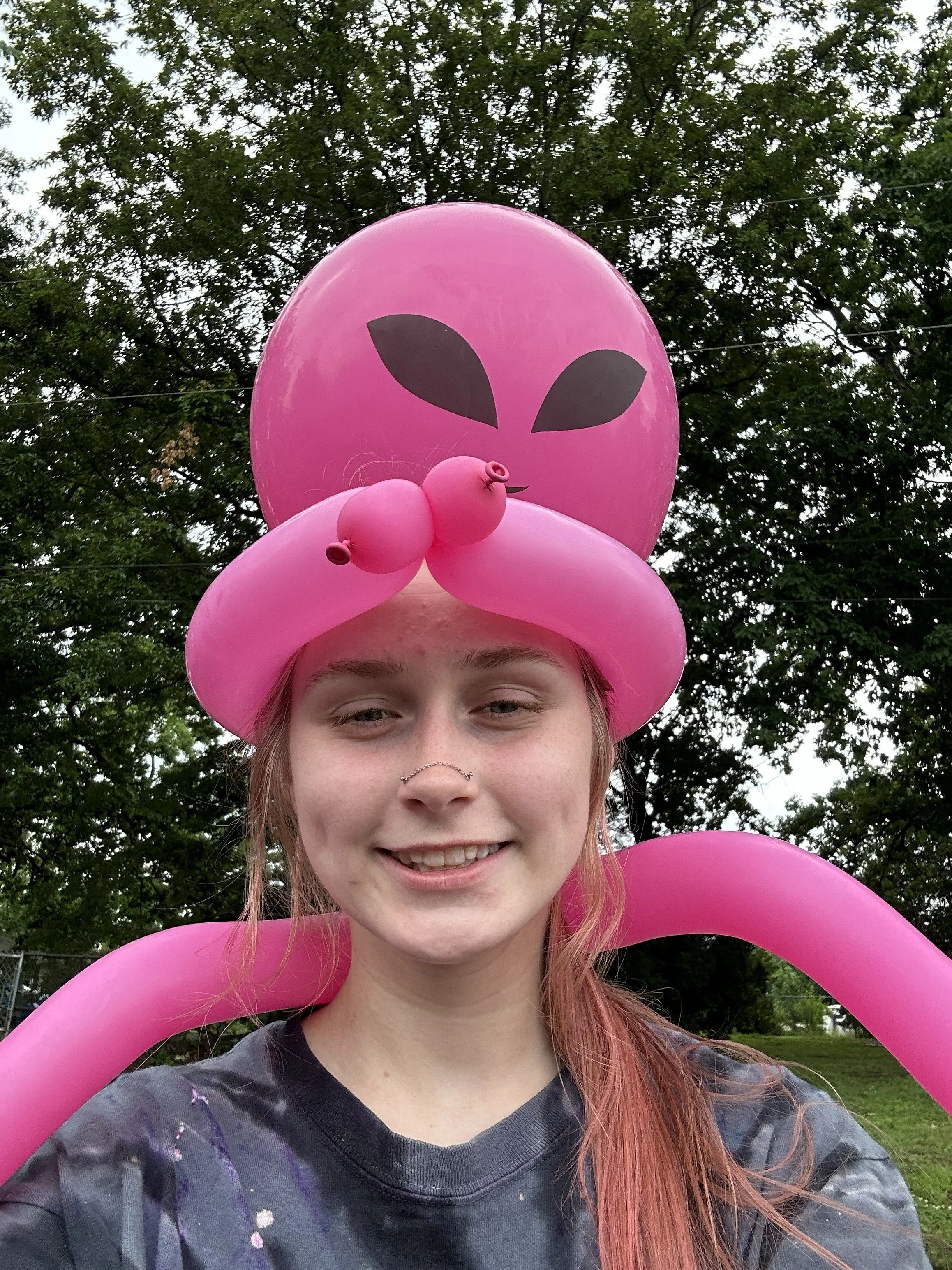 A young person with pink hair and a septum piercing smiling outdoors, wearing a black tie-dye shirt, surrounded by pink balloon decorations, with trees in the background.