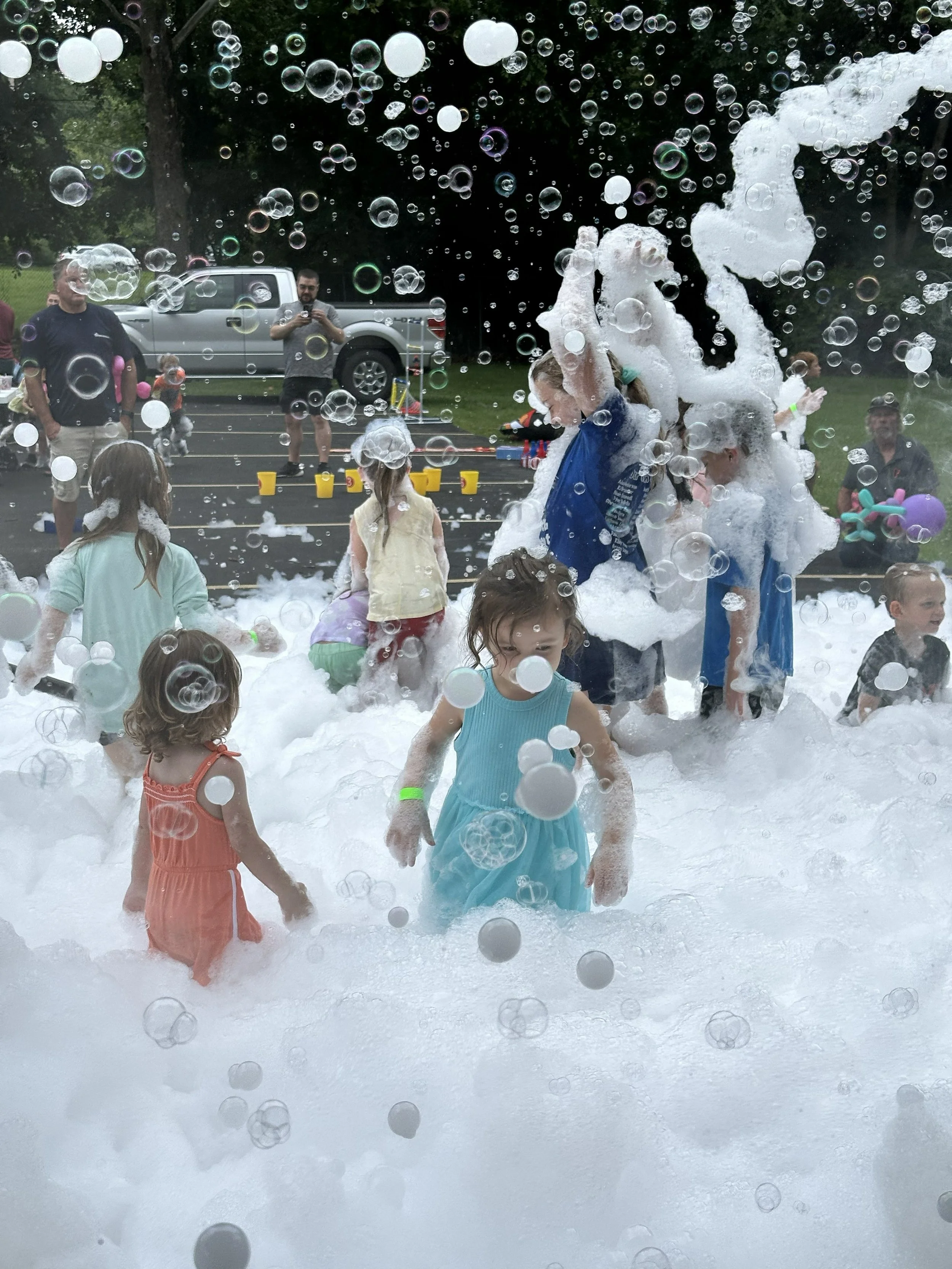 Kids and adults playing in a foam and bubble-filled outdoor area, with some people taking photos and a truck in the background.