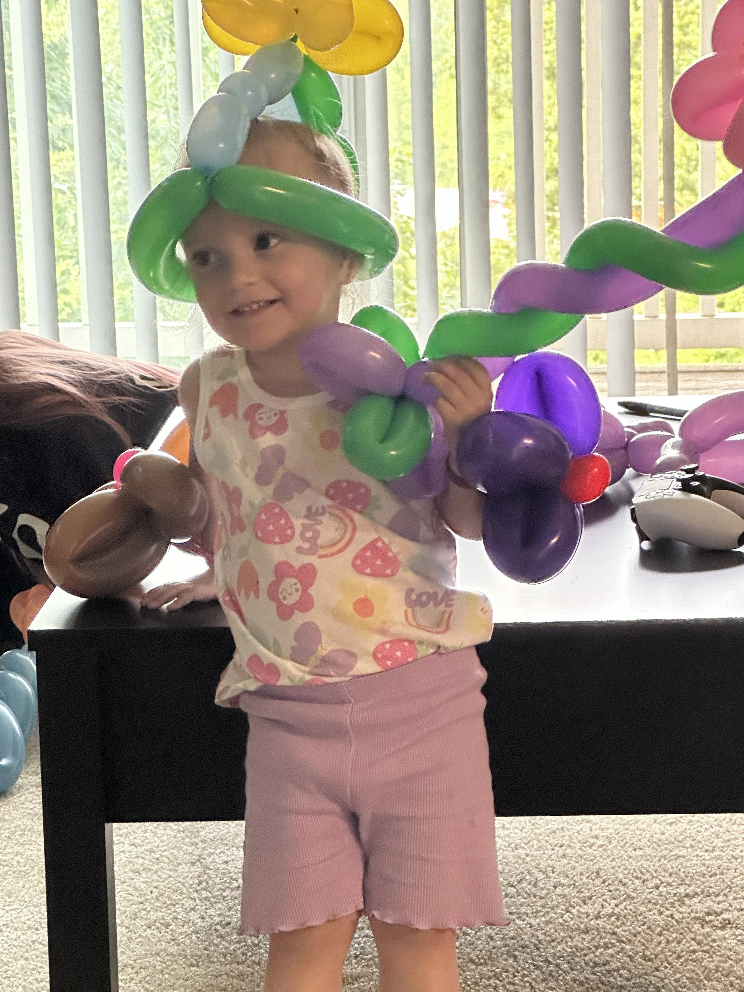 A young girl wearing a balloon hat and holding colorful balloon decorations indoors near a window with vertical blinds.