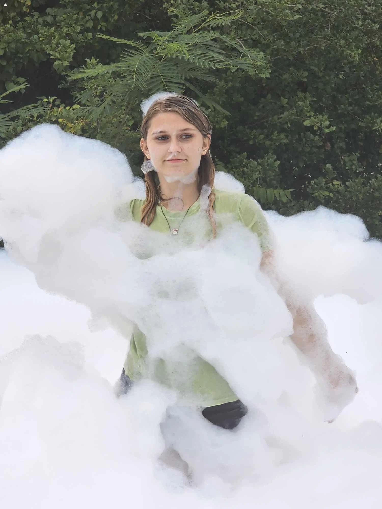 A woman with wet hair and a green shirt sitting in a bubble bath outdoors with green foliage in the background.