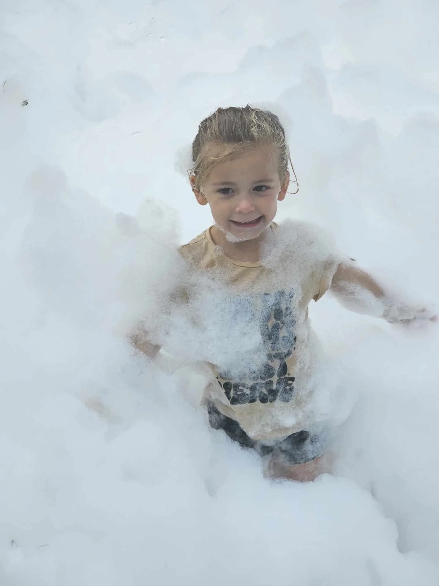 A young girl with wet blonde hair and a big smile standing in a bubble bath with white foam all around him.
