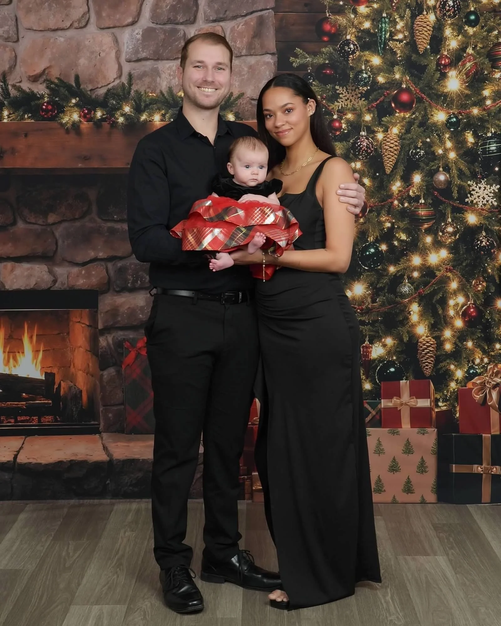 A family of three posing in front of a Christmas tree decorated with ornaments and lights, with a fireplace, stockings, and wrapped presents nearby.