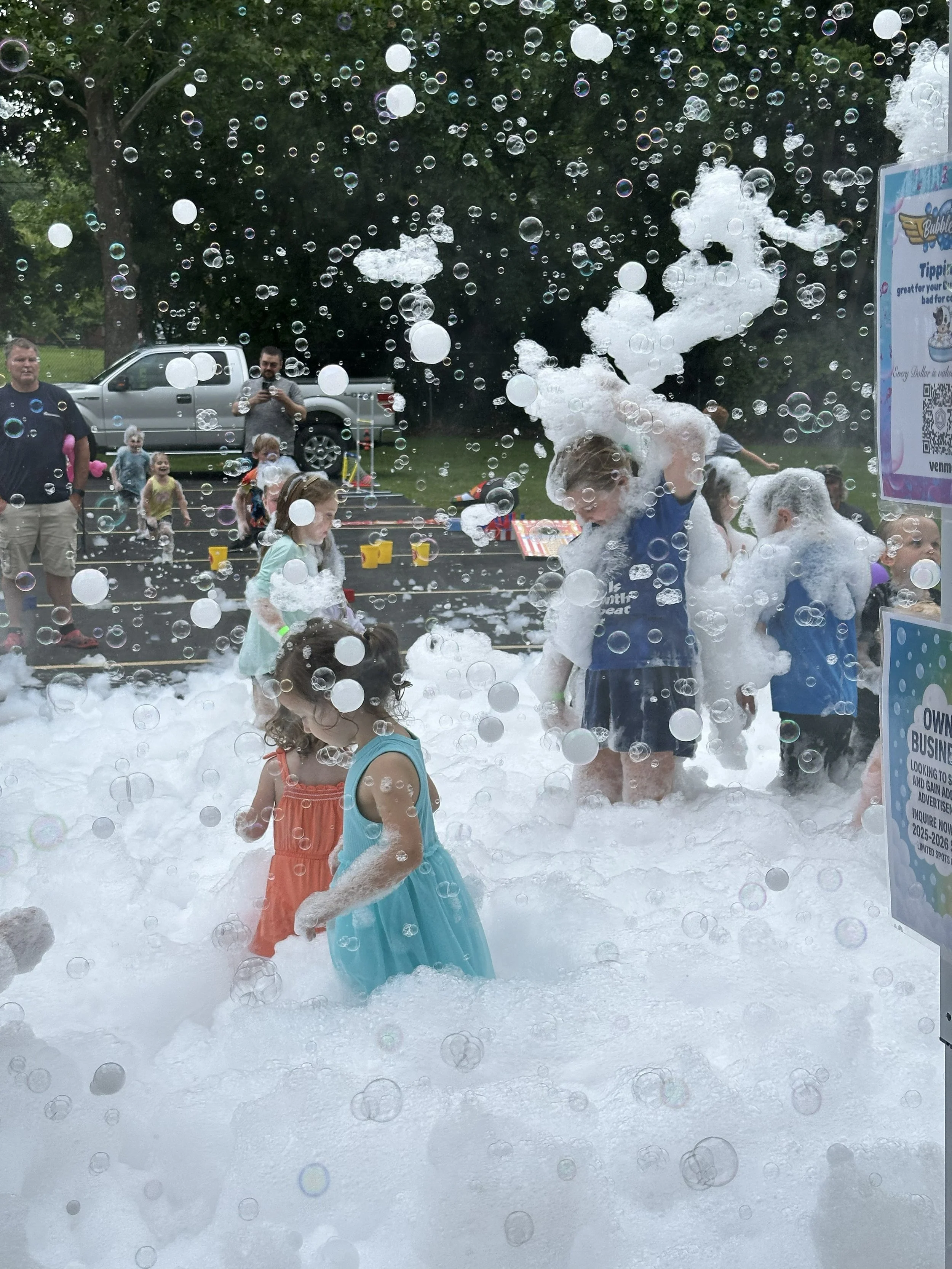 Children playing in foam at an outdoor event, with bubbles floating in the air and adults observing. Trees and a vehicle are visible in the background.