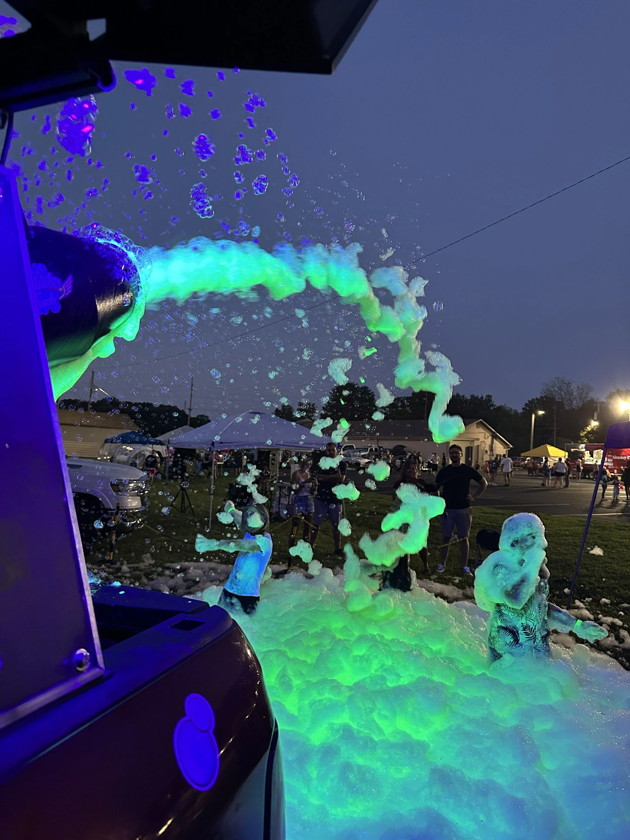 People enjoying a foam party outdoors at night with foam and bubbles illuminated by colorful lights, with tents and cars in the background.