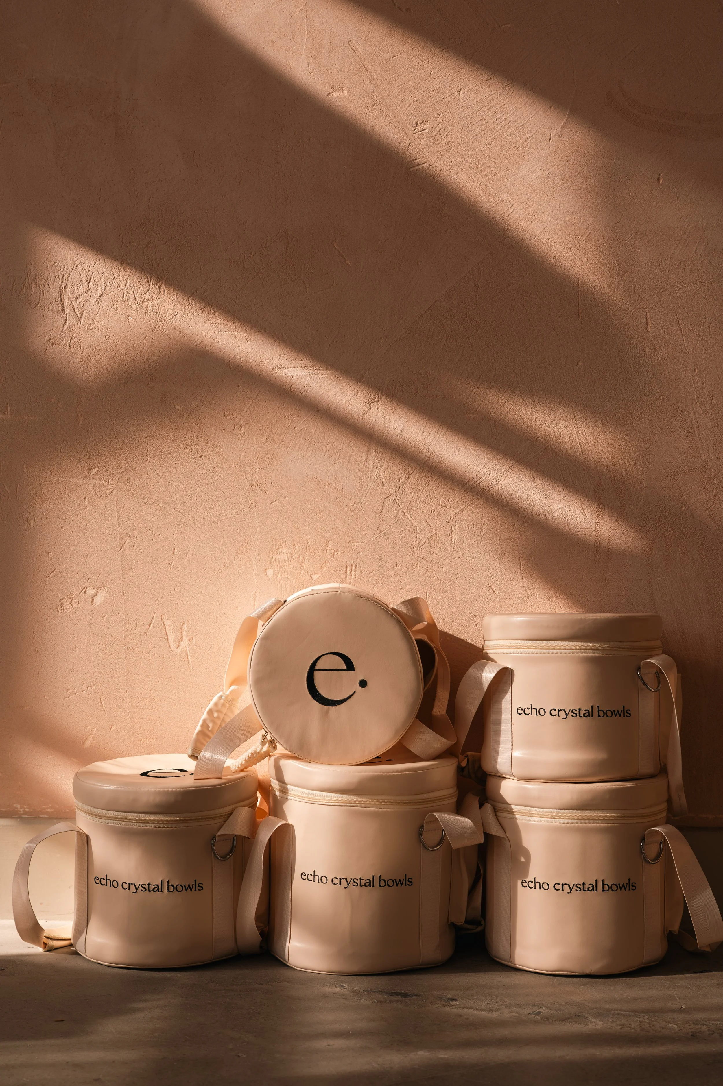 Multiple cream-colored echo crystal bowls with beige ribbons and tags that read "echo crystal bowls," placed on a concrete floor against a textured, pinkish-beige wall with shadows cast from above.