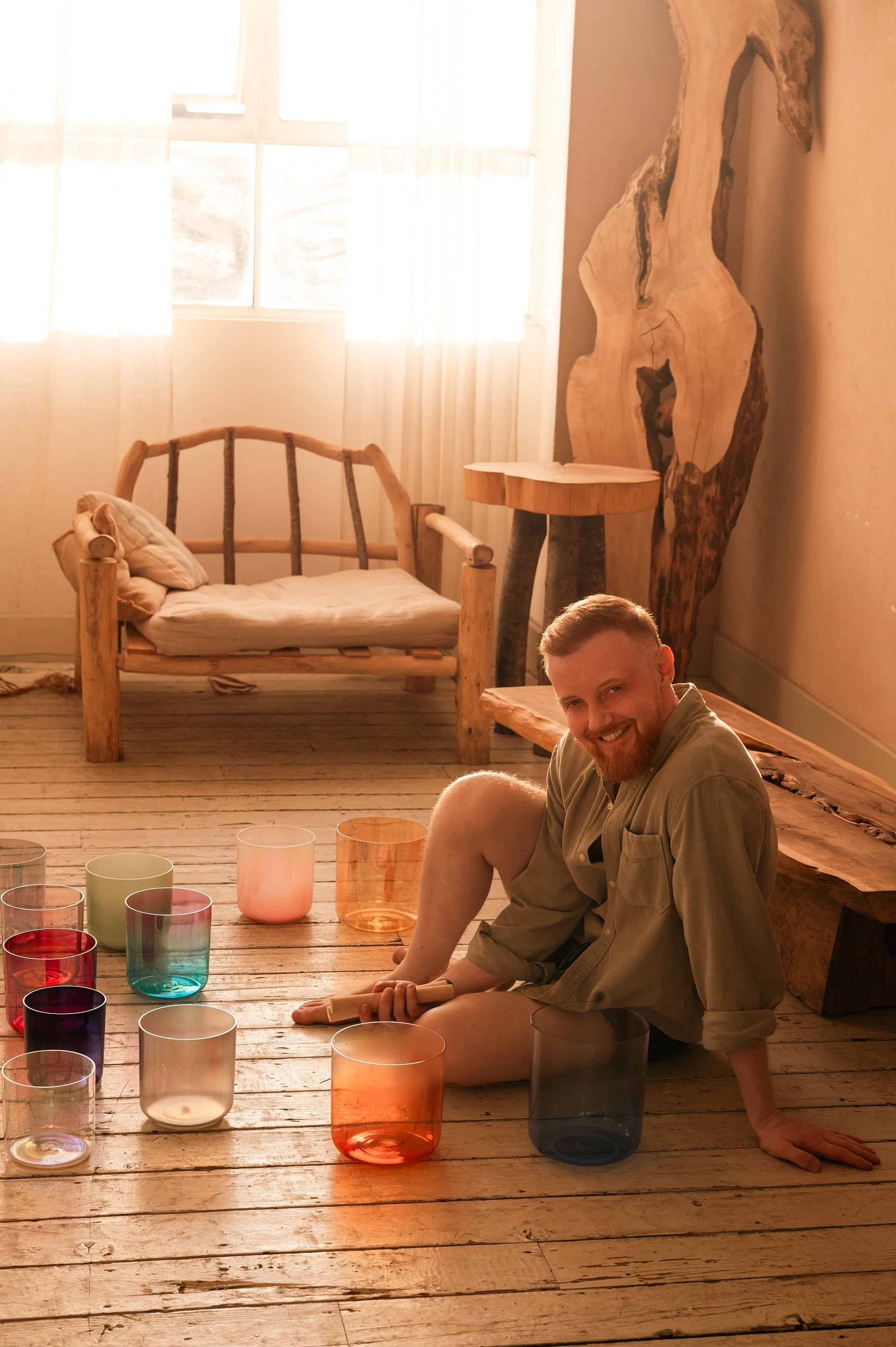 A man with a beard sitting on the floor in a room with wooden floors and furniture, surrounded by colorful glass cups, with sunlight coming through a window and a large natural wood art piece on the wall.