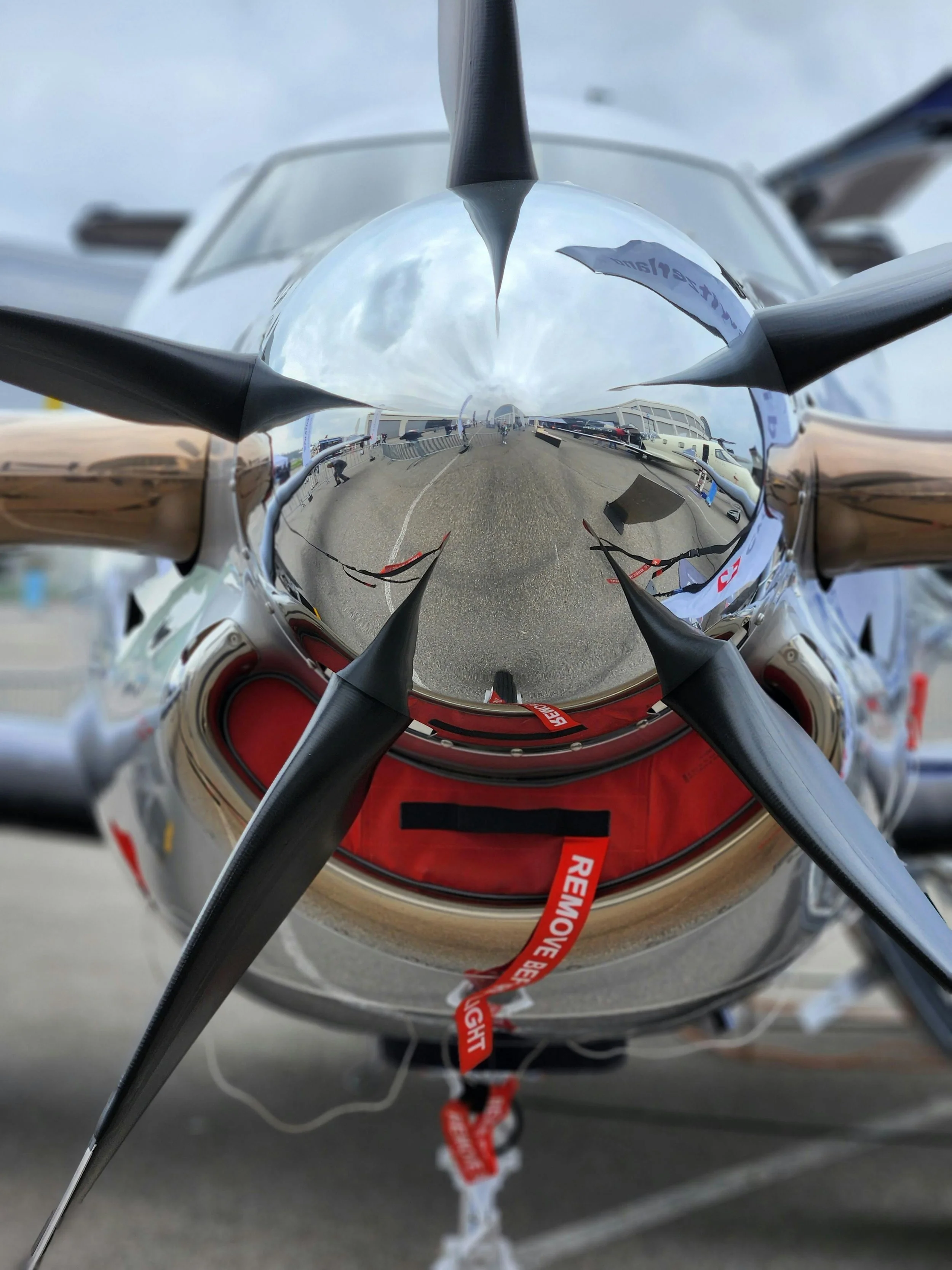 A close-up view of a reflective, metallic boat propeller with several blades, attached to a boat, with a cloudy sky and boats/reflections visible in the surface.