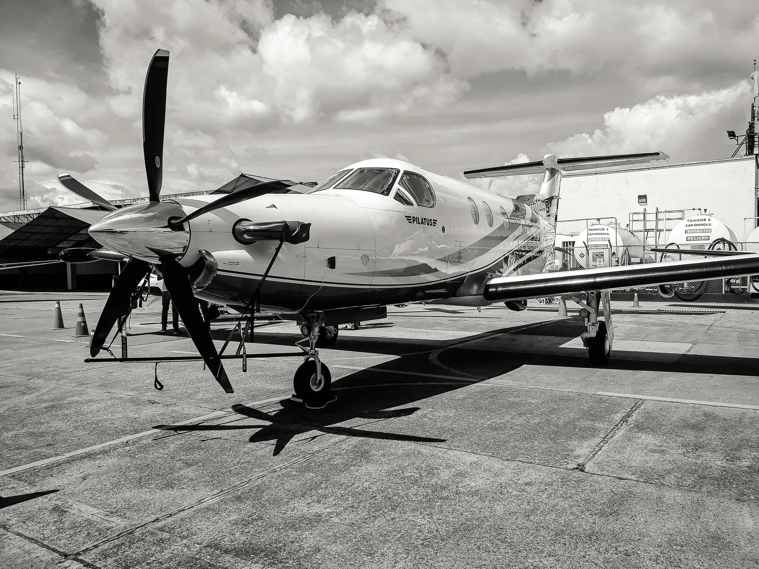A small private jet parked on an airport tarmac with fuel tanks and industrial equipment nearby, under cloudy skies.