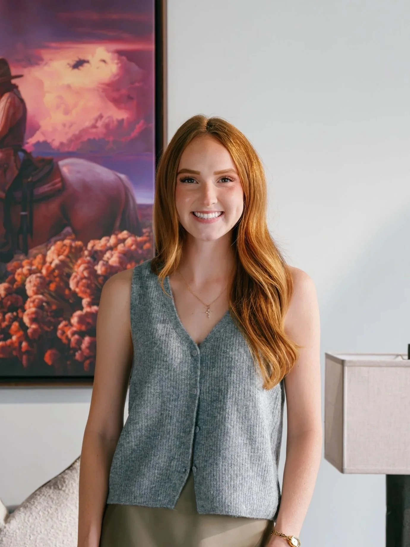 A young woman with long red hair, smiling, wearing a gray sleeveless button-up vest and a beige skirt, standing indoors in front of a large colorful painting of a horse and a woman, and a modern white wall.