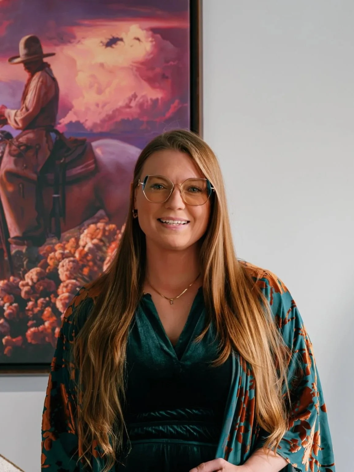 A woman with long hair, glasses, and a colorful patterned shirt standing in front of a vibrant painting of a cowboy riding a horse against a sunset sky.