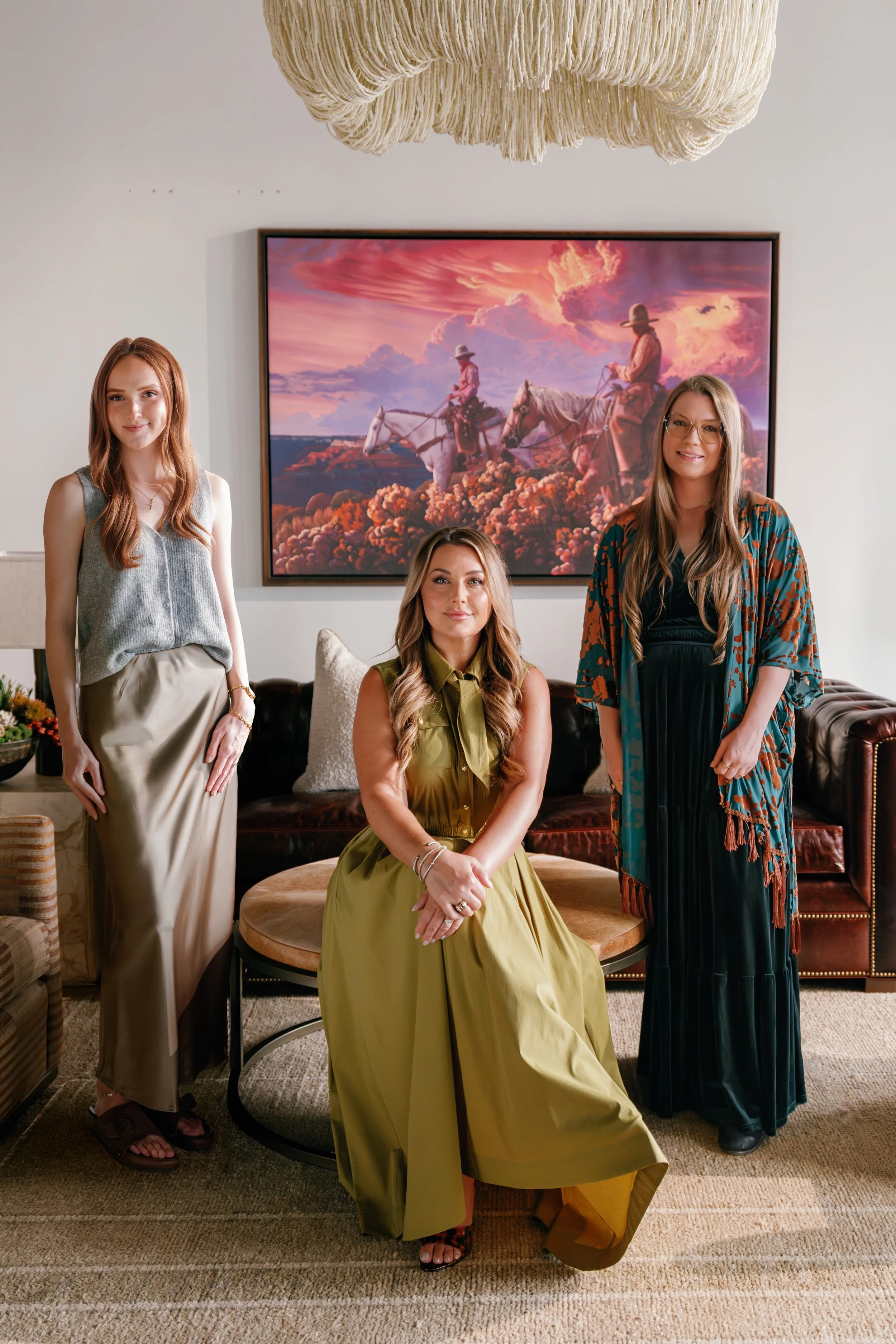 Three women posing in a living room in front of a wall-mounted painting of two women riding horses during a colorful sunset. One woman is seated on a round ottoman, wearing a yellow-green dress, while the other two women are standing, one in a gray sleeveless top and beige skirt, and the other in a patterned kimono-style robe over a black dress.