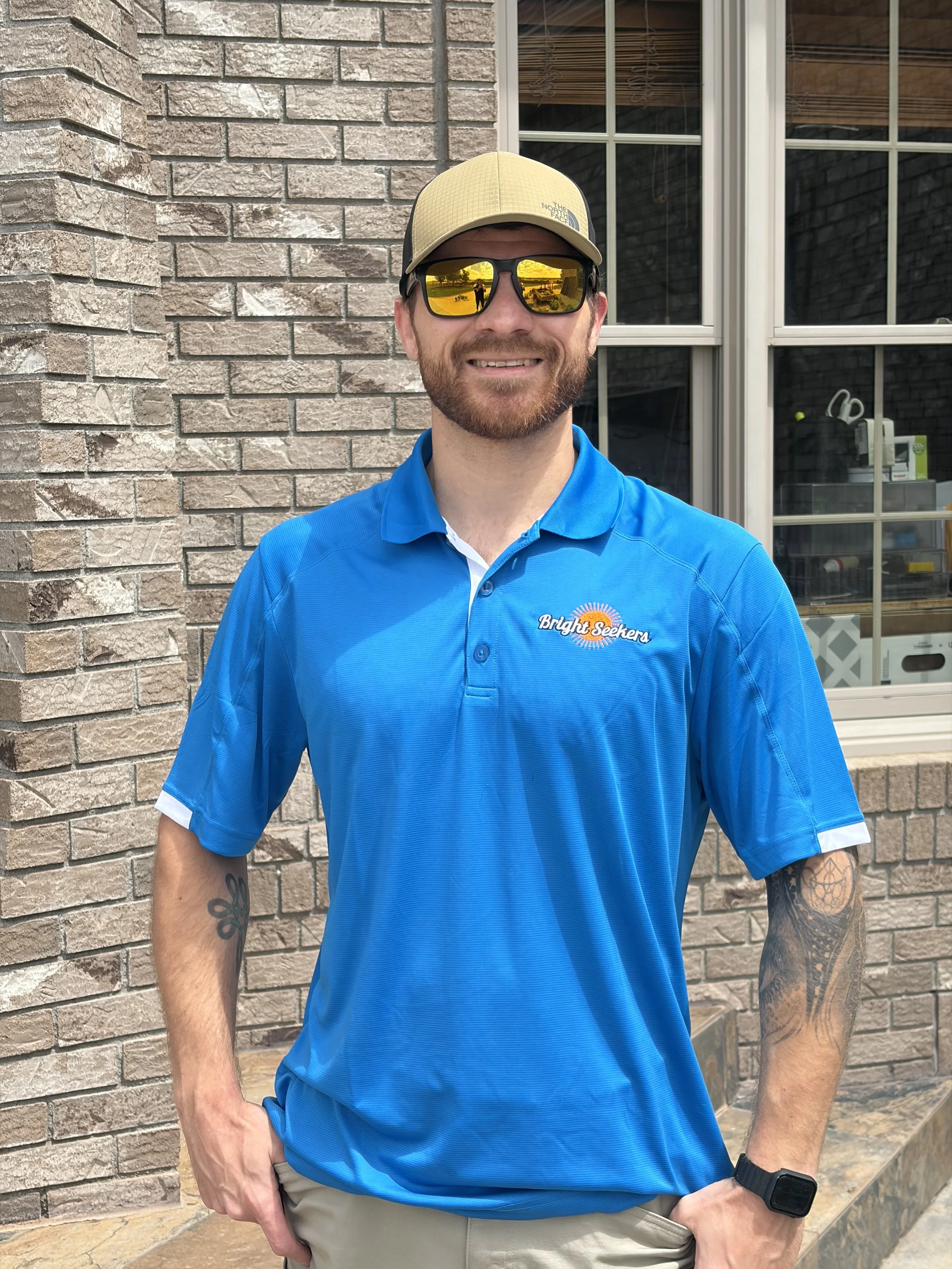 A man with a beard wearing a beige cap, reflective sunglasses, a blue polo shirt with 'Bright Seekers' logo, and beige pants. He is standing outdoors in front of a brick wall and window, with one hand in his pocket and the other resting on his hip.