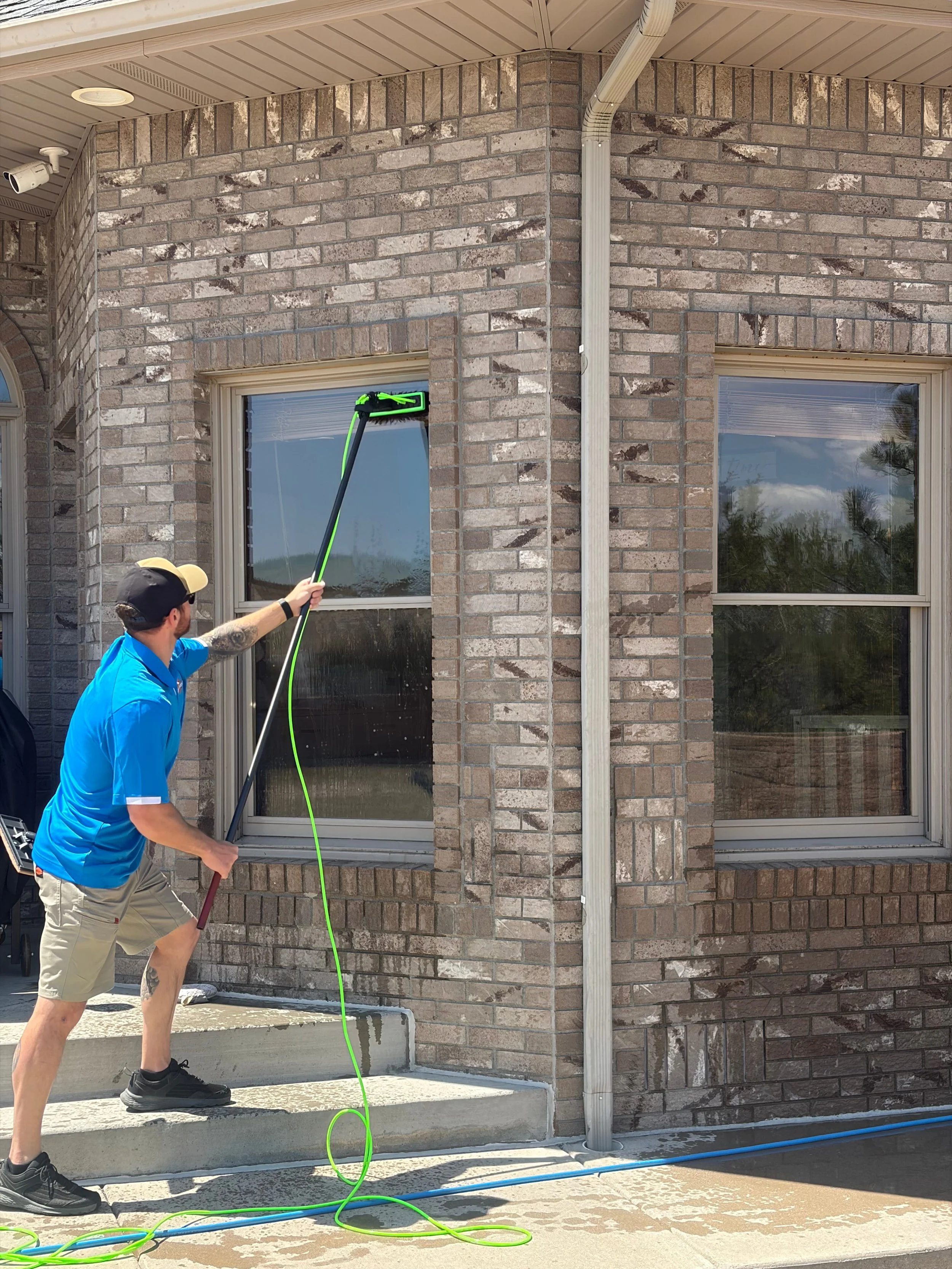 A man cleaning the exterior window of a brick house with a long-handled squeegee, standing on the concrete step of the house's porch.