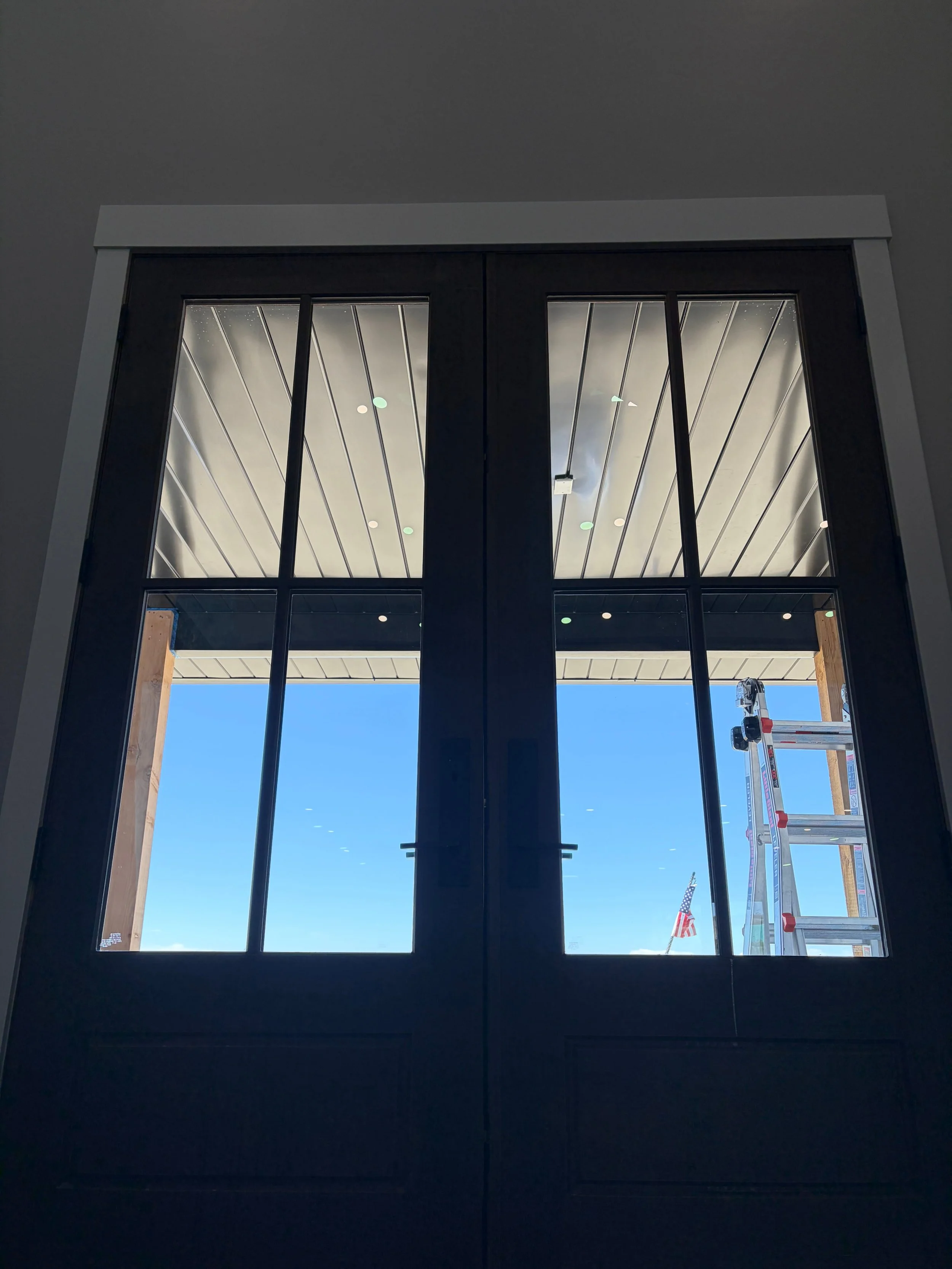 Photo of interior black-framed glass double doors looking outside to a clear blue sky, with a ladder and American flags visible through the glass.