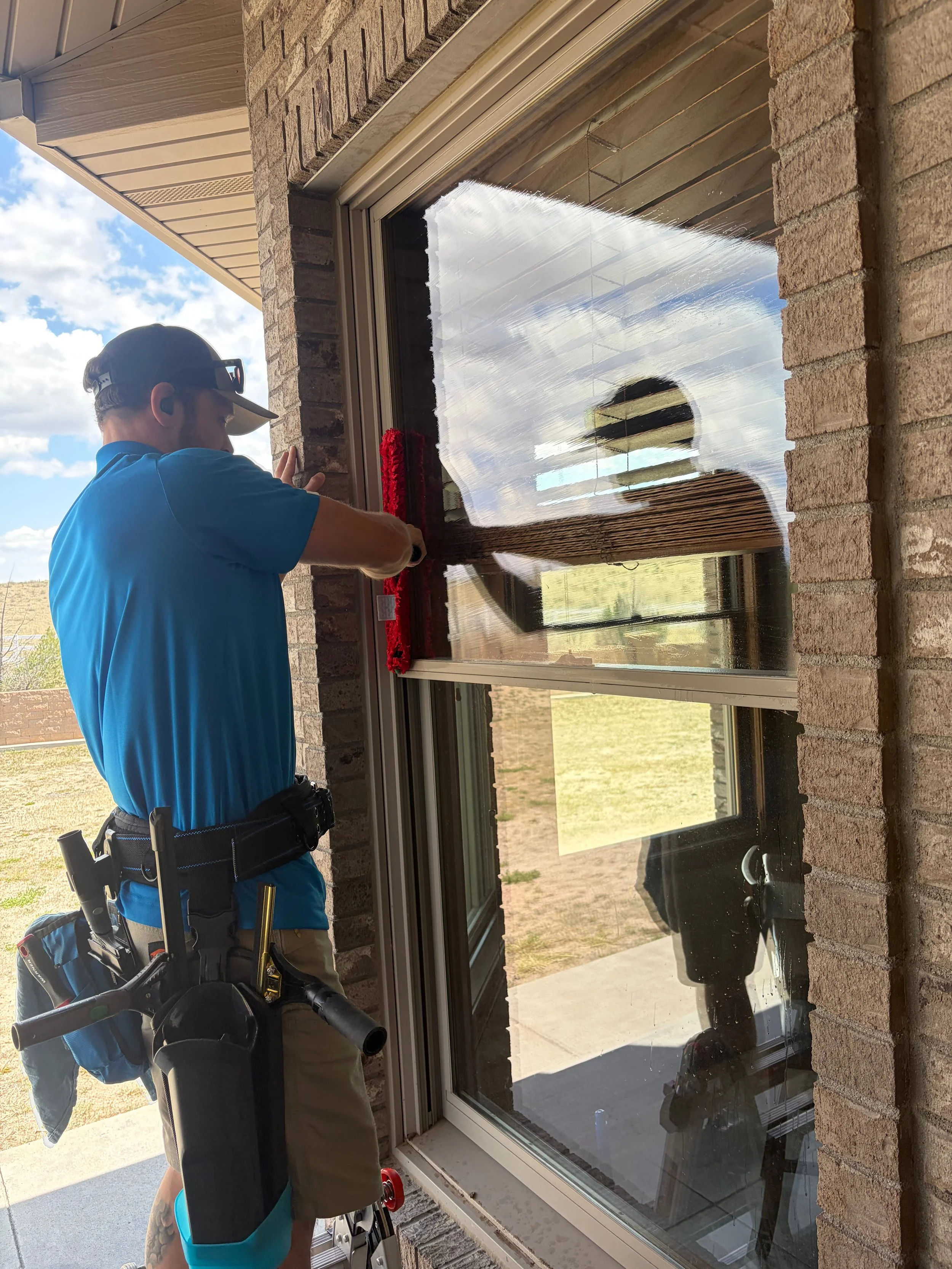 A person in a blue shirt and tan pants, with a tool belt, using a tool to apply a red decorative item to a window. They are outside a brick house. The reflection of the person can be seen in the window, with a cloudy sky in the background.