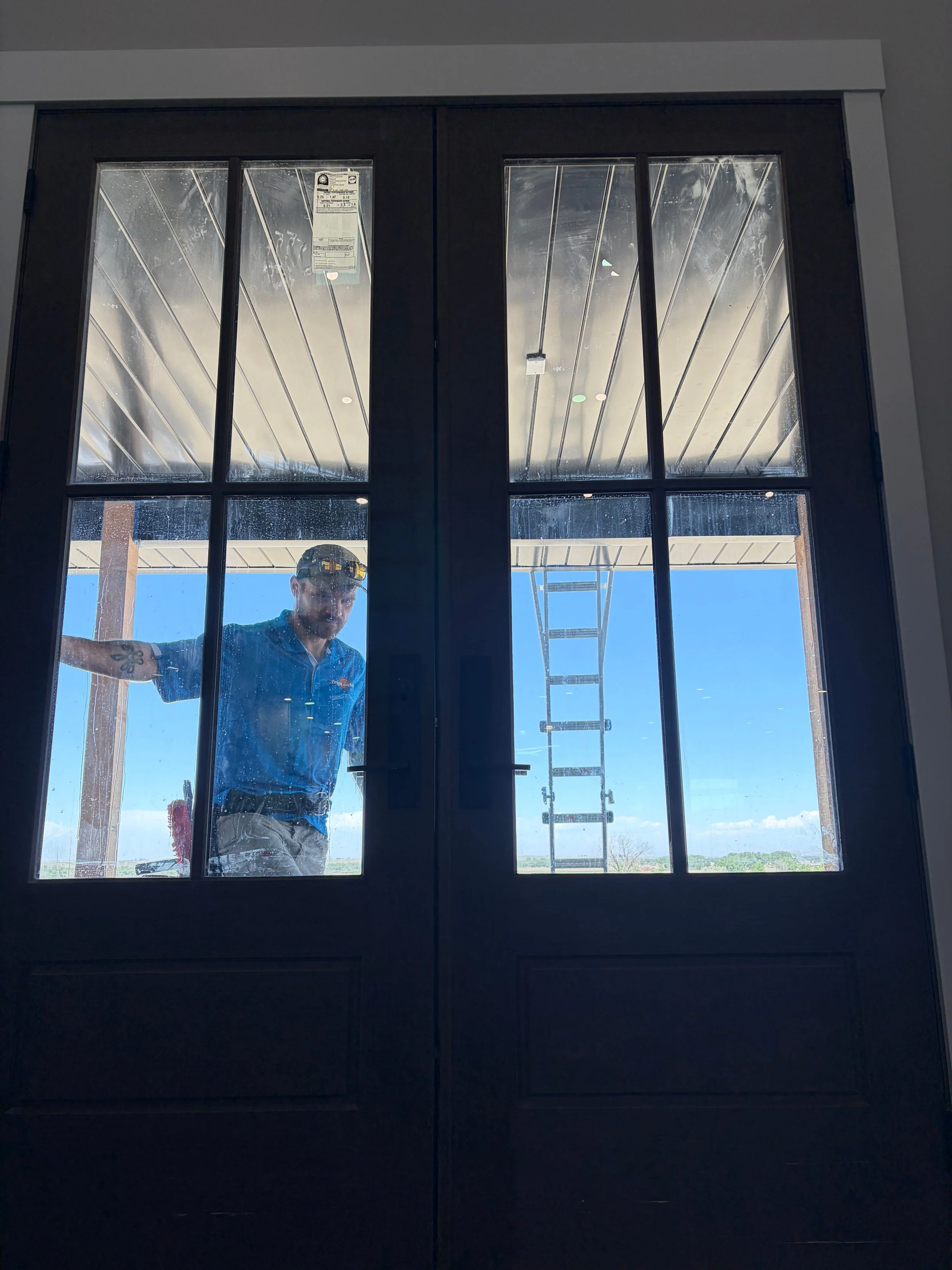 A man is cleaning a glass door, seen from inside a building, with sunlight and a blue sky visible outside. The man is wearing safety glasses and a blue shirt.