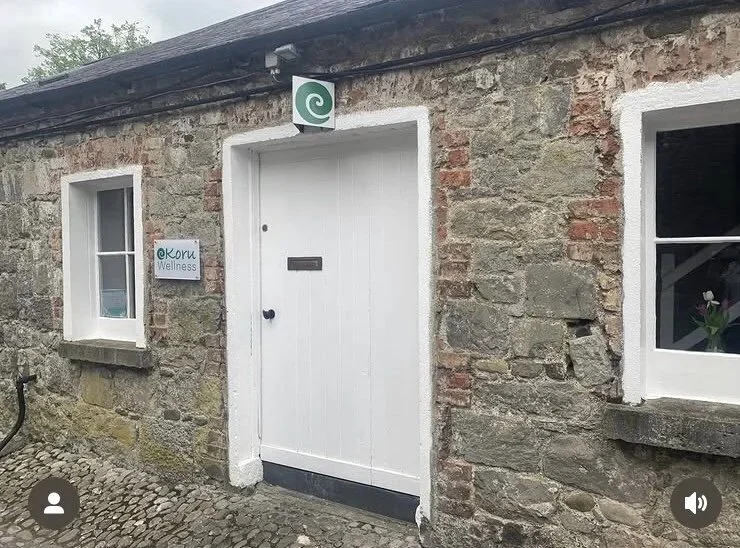 Stone building with white door and two small windows; one window has a flower inside. A sign next to the door reads 'eKoru Wellness.' Above the door, there is a spiral sign, likely indicating a wellness or holistic center.