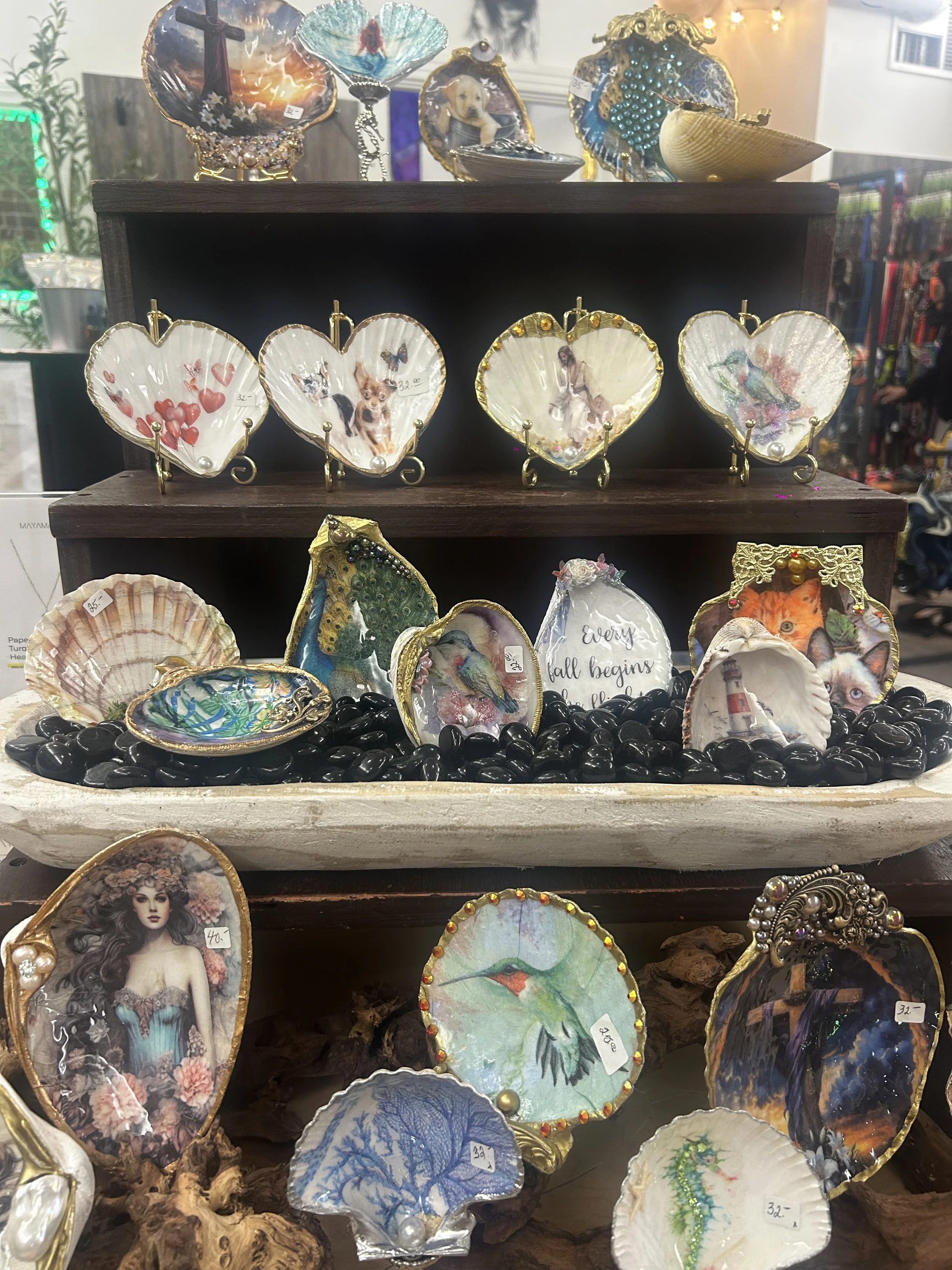 Decorative shells with various artistic designs and illustrations displayed on a wooden shelf, some with photographs and ornaments inside.