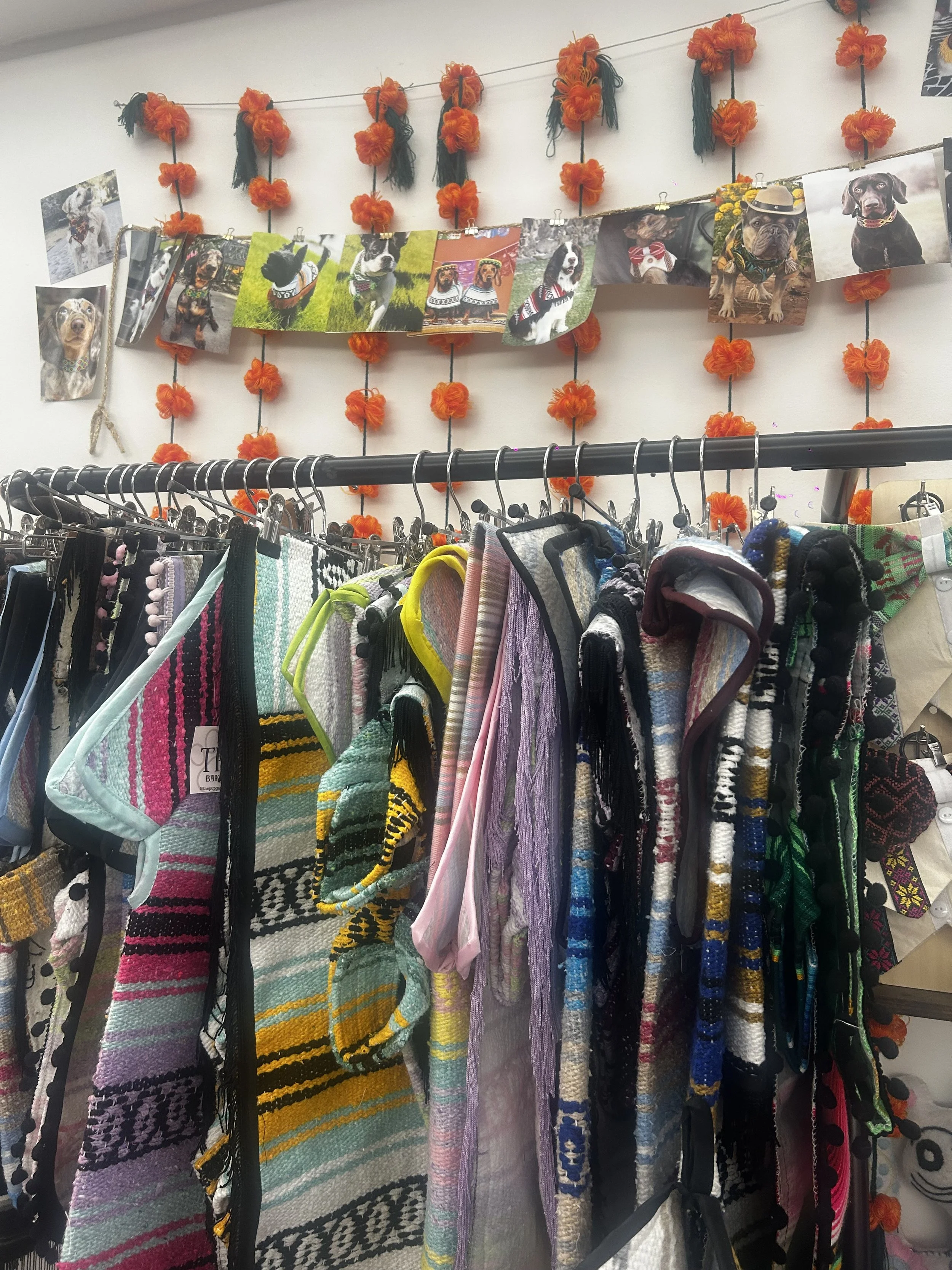 Clothing rack with colorful woven bags and scarves, dog photos and orange floral decorations hanging on the wall behind.