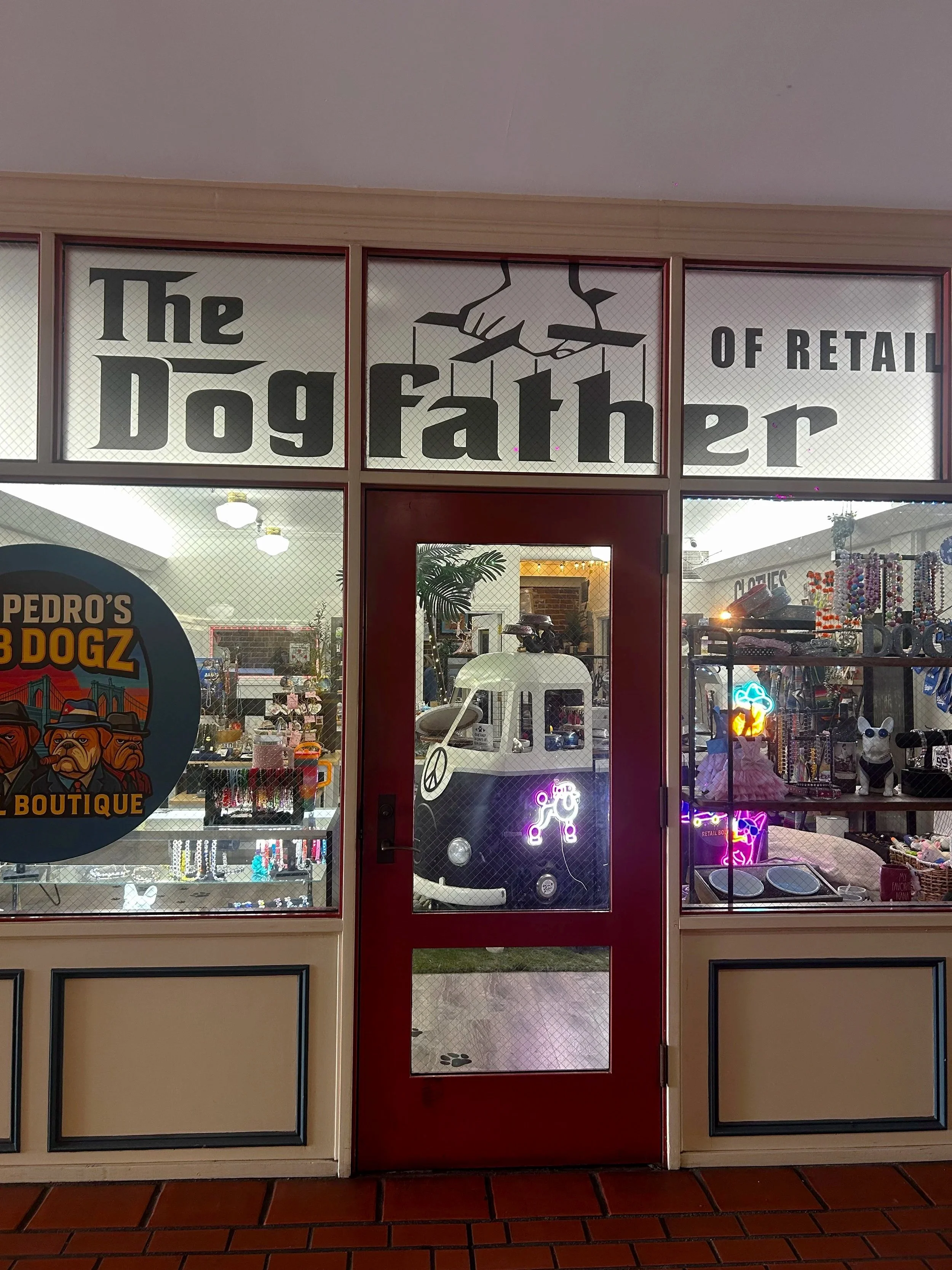 Storefront window with signage that reads "The Dog Father of Retail" and a graphic of a hand holding marionette strings. The storefront displays various pet accessories and neon light decorations inside.