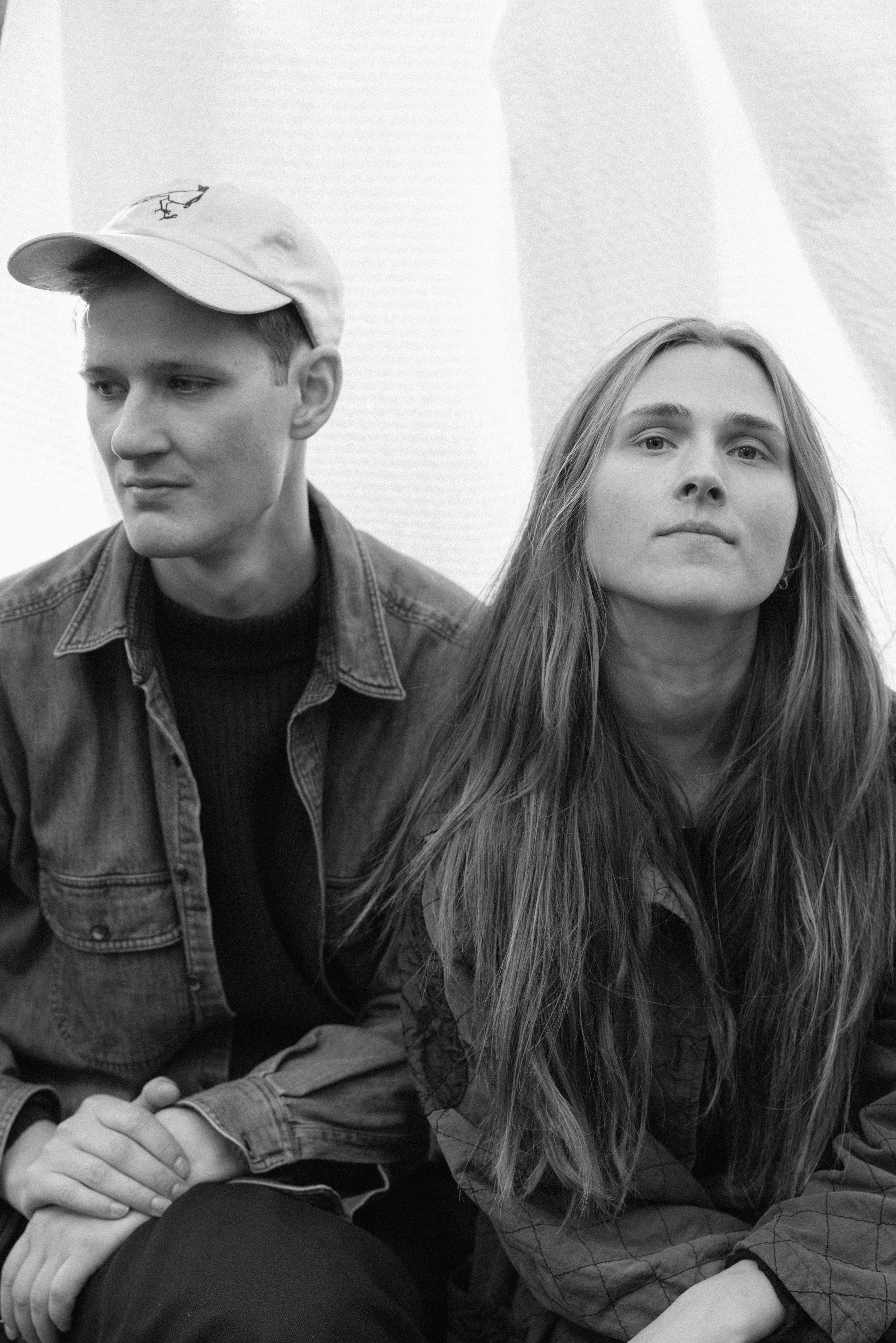 Black and white photograph of young man and woman sitting together. The man is wearing a baseball cap with a small dinosaur embroidered on it, a denim jacket, and a black shirt. The woman has long hair and is wearing a checkered shirt. Both appear serious and are looking in different directions.