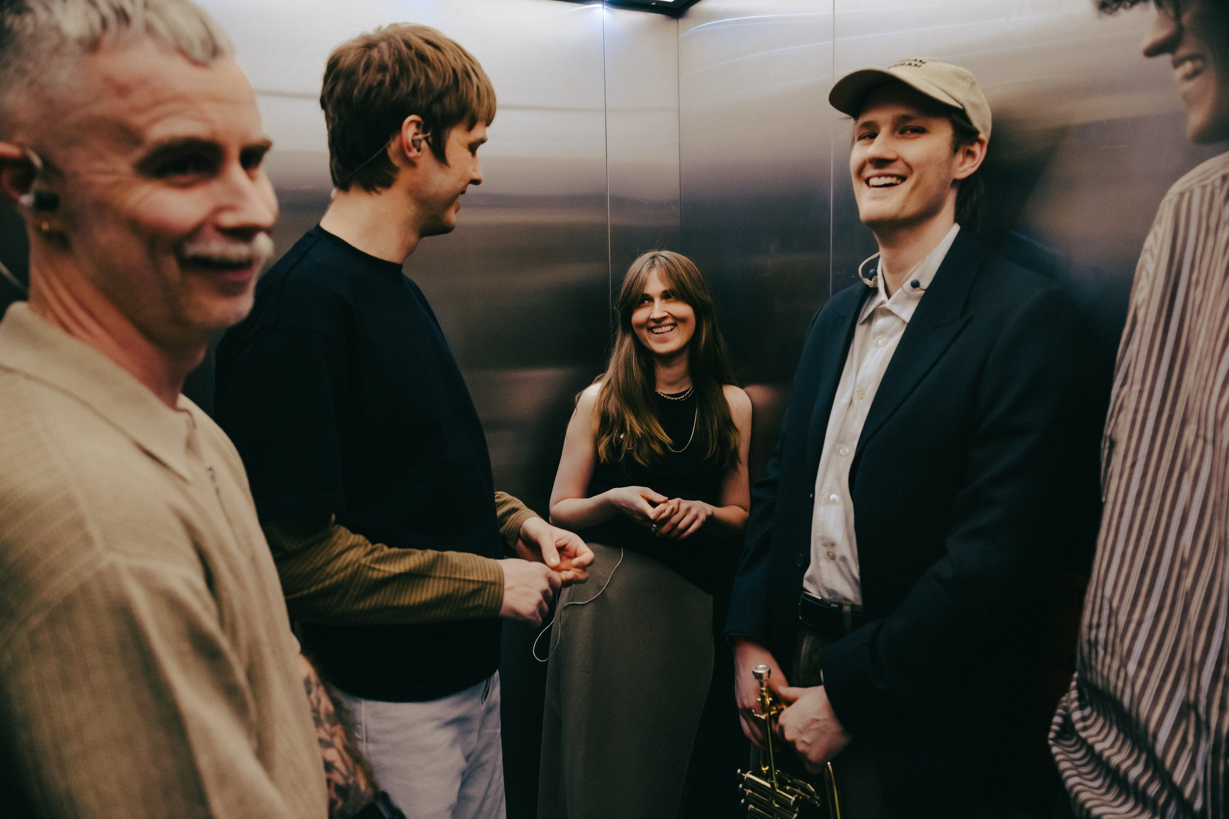Five people in an elevator, two men shaking hands, and smiling, with a woman observing in the background.