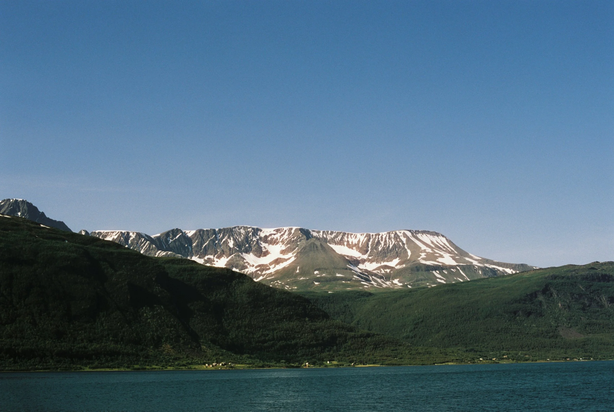 Scenic view of snow-capped mountains across a lush green landscape and a calm lake with clear blue sky