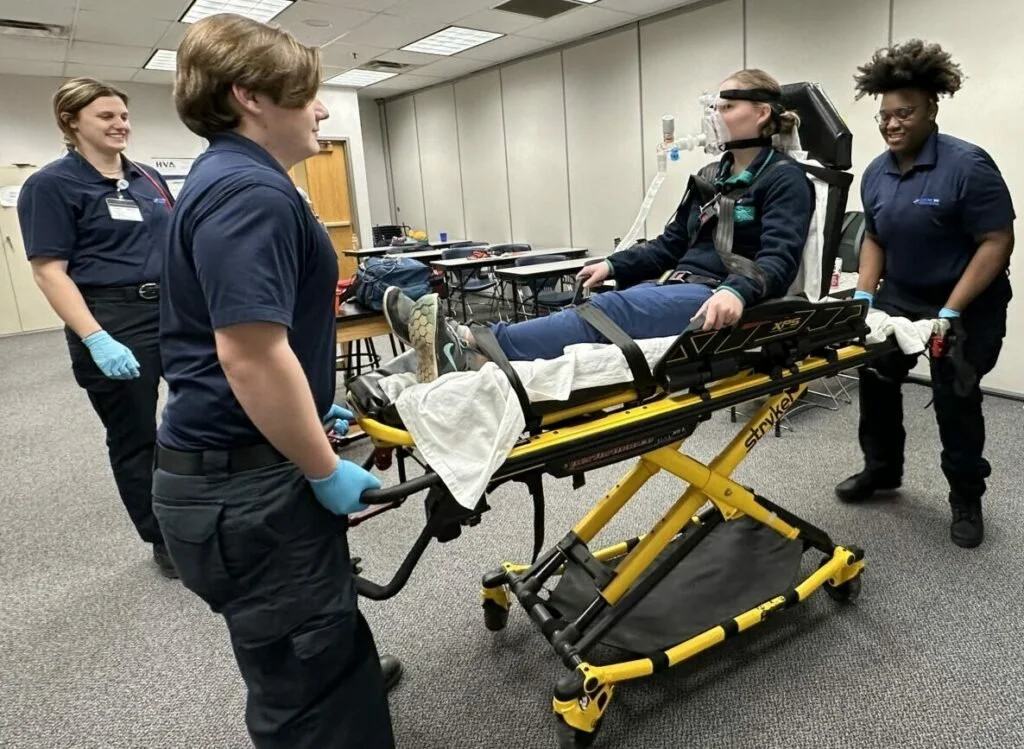 Four EMS/EMT students, two women and two men, are assisting a patient sitting on a stretcher in a sim lab room. The patient is wearing a headgear connected to breathing apparatus. The professionals are  wearing gloves, ready to help.