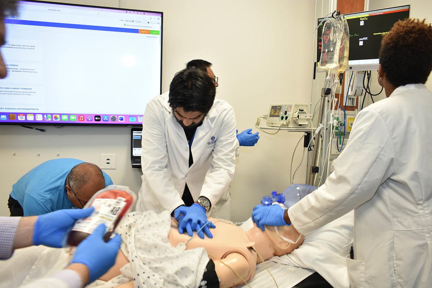 Medical students performing a procedure on a patient mannequin in a clinical setting, with monitors and medical equipment in the background.